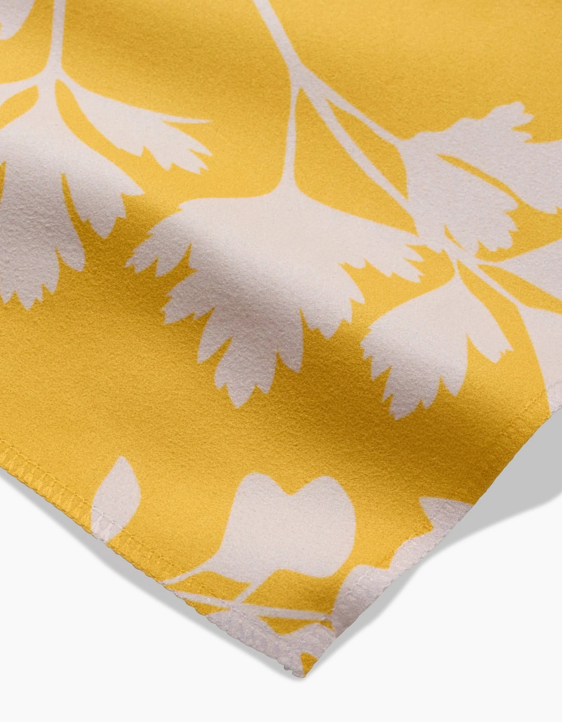 A close-up of GEOMETRY’s Garden of Gold Geosuede™ fabric shows white botanical leaves and detailed edge stitching. Made from recycled materials, these Dinner Napkins bring style and sustainability to your table.