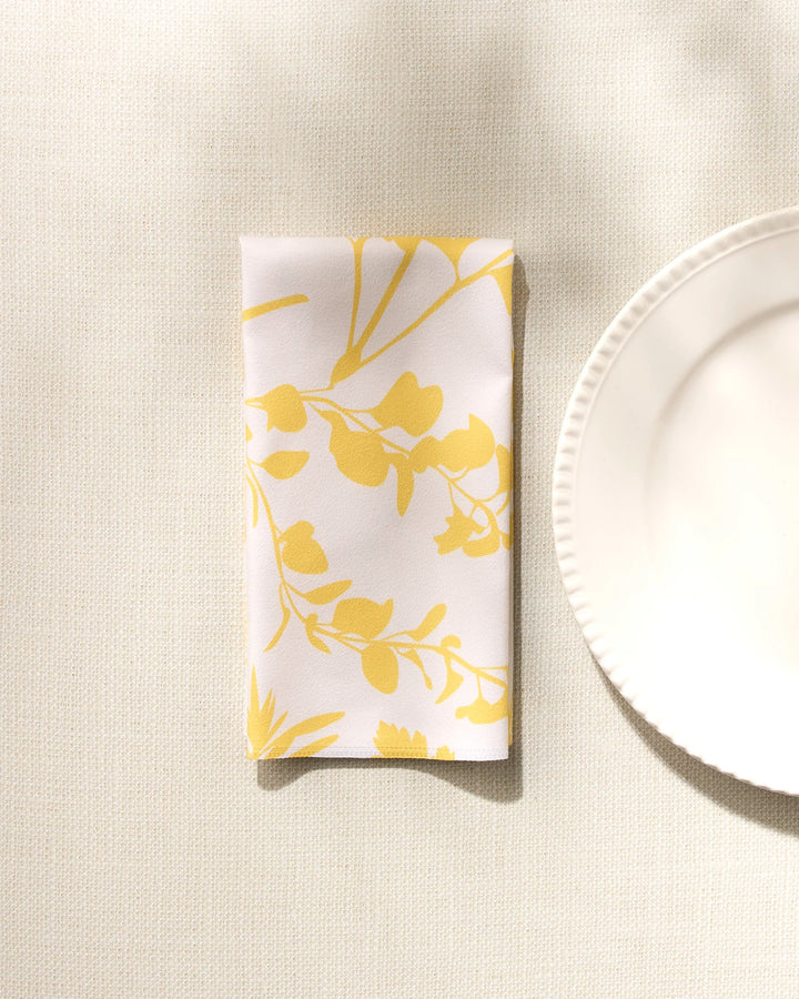 The GEOMETRY "Garden of Gold" folded white washable cloth napkin, featuring a yellow floral pattern, rests beside a scalloped-edge white plate on a light textured surface under soft natural light.
