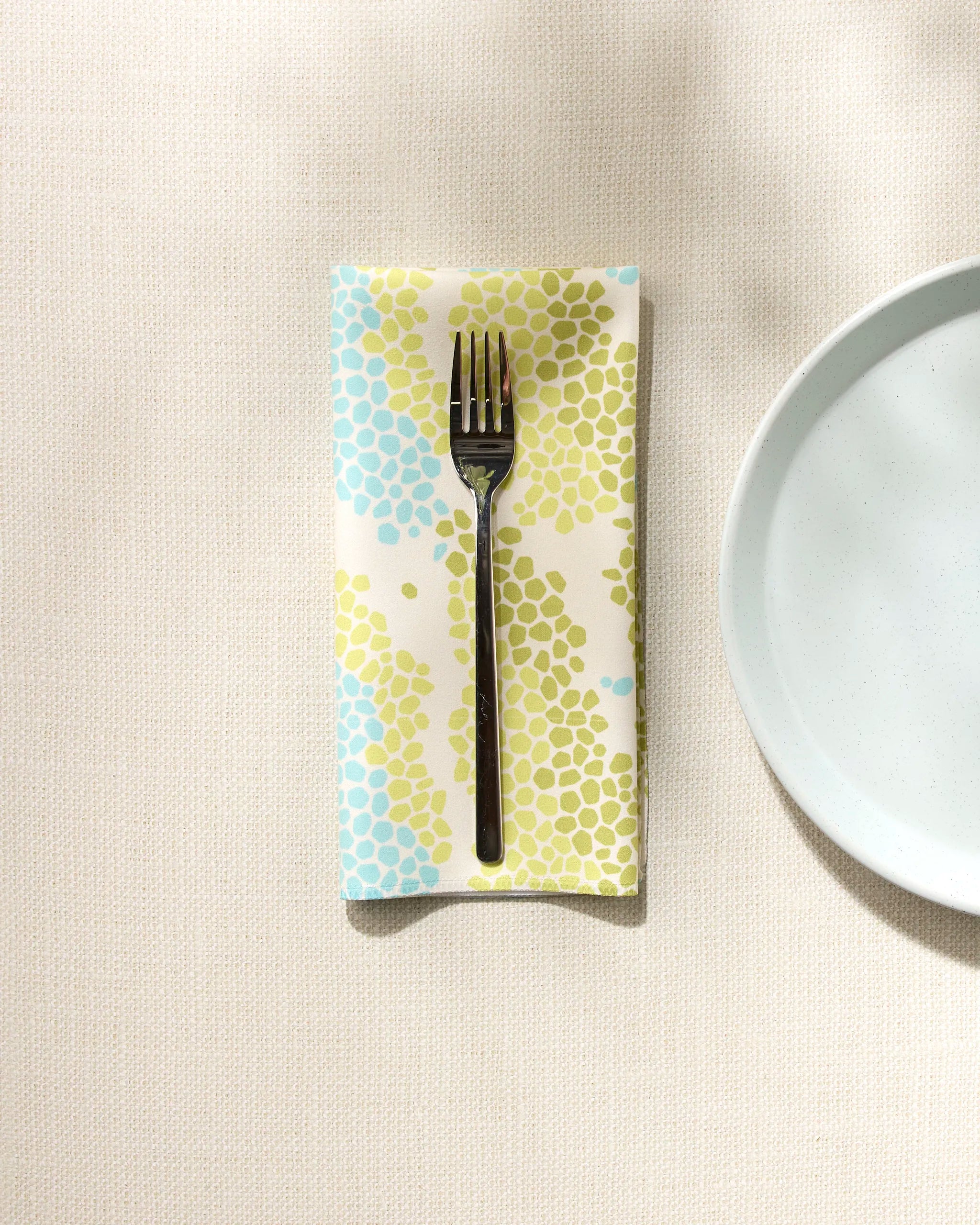 A Geo Shell napkin by GEOMETRY with blue and green dots rests under a fork on a cream tablecloth, beside a plain white plate.
