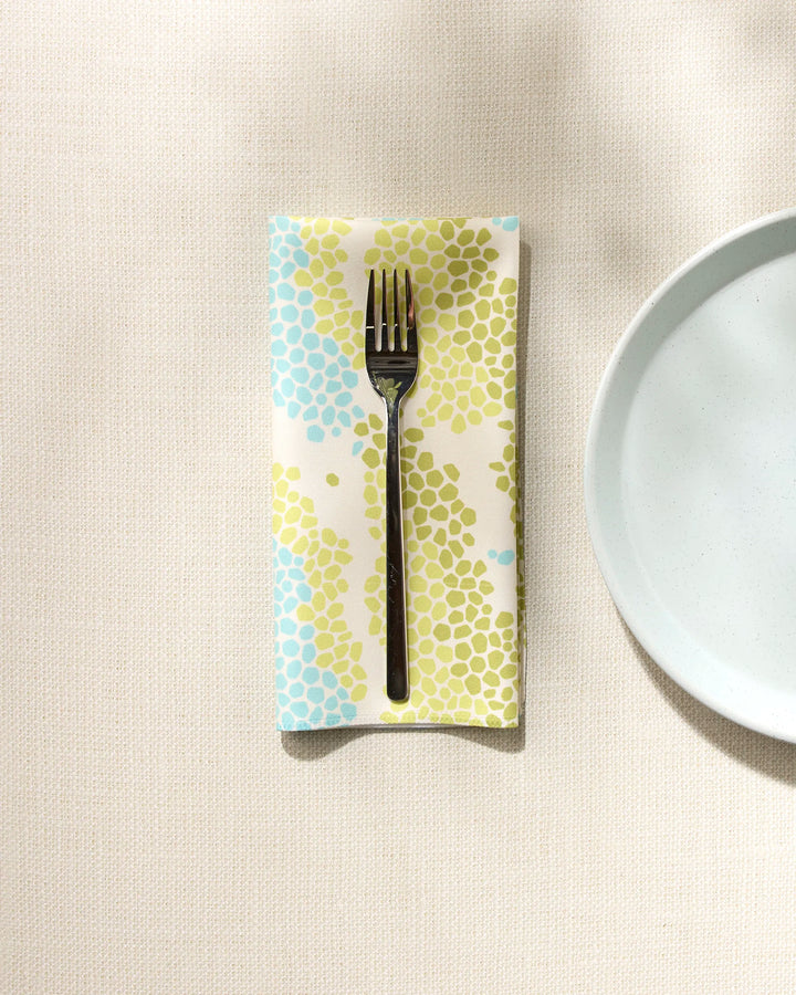 A Geo Shell napkin by GEOMETRY with blue and green dots rests under a fork on a cream tablecloth, beside a plain white plate.