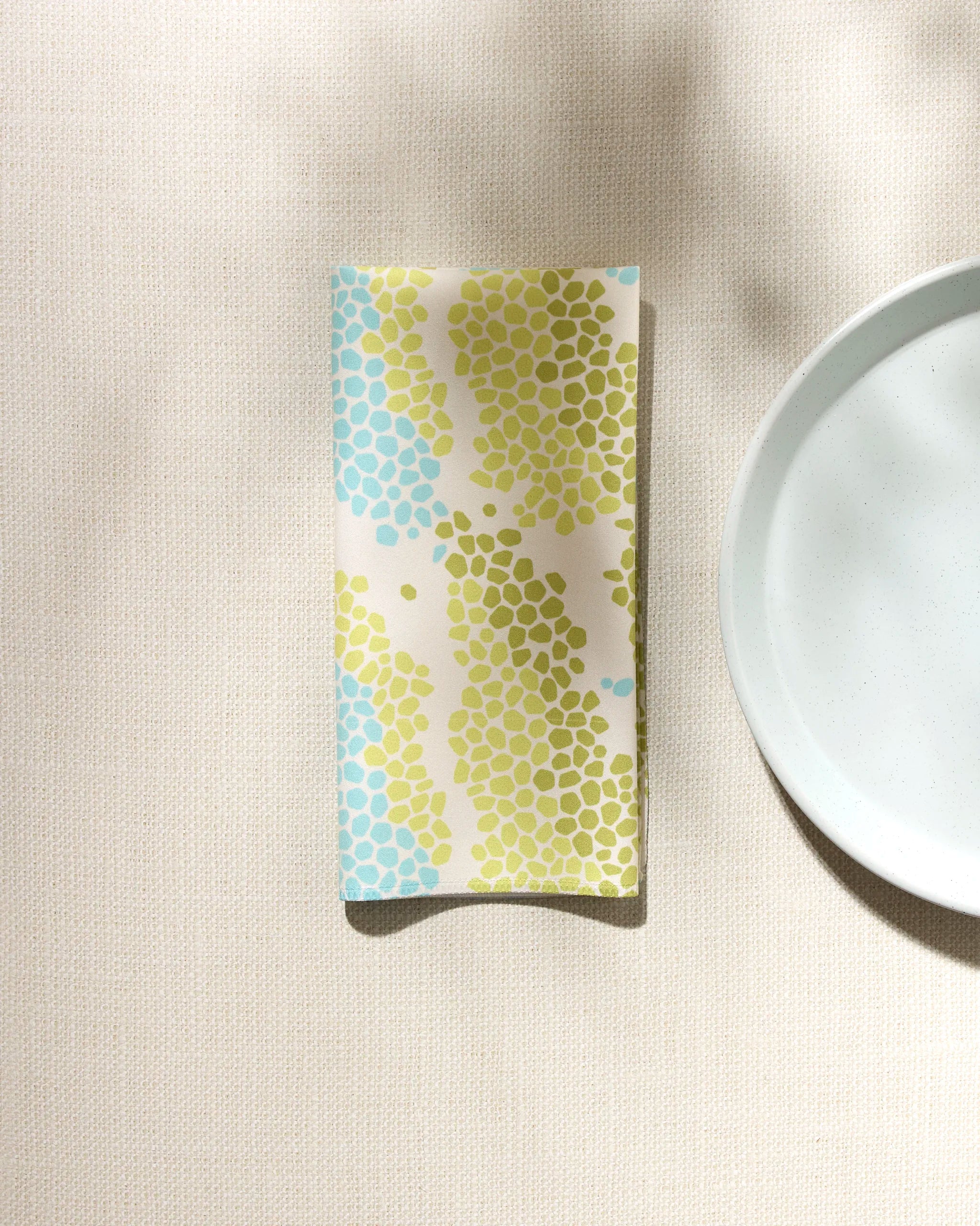 The GEOMETRY Geo Shell absorbent cloth napkin, featuring blue and green hexagonal patterns, rests beside a plain white plate on a light textured surface with soft shadows.