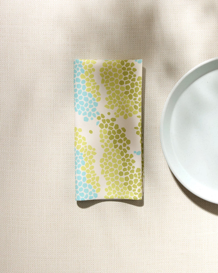 The GEOMETRY Geo Shell absorbent cloth napkin, featuring blue and green hexagonal patterns, rests beside a plain white plate on a light textured surface with soft shadows.