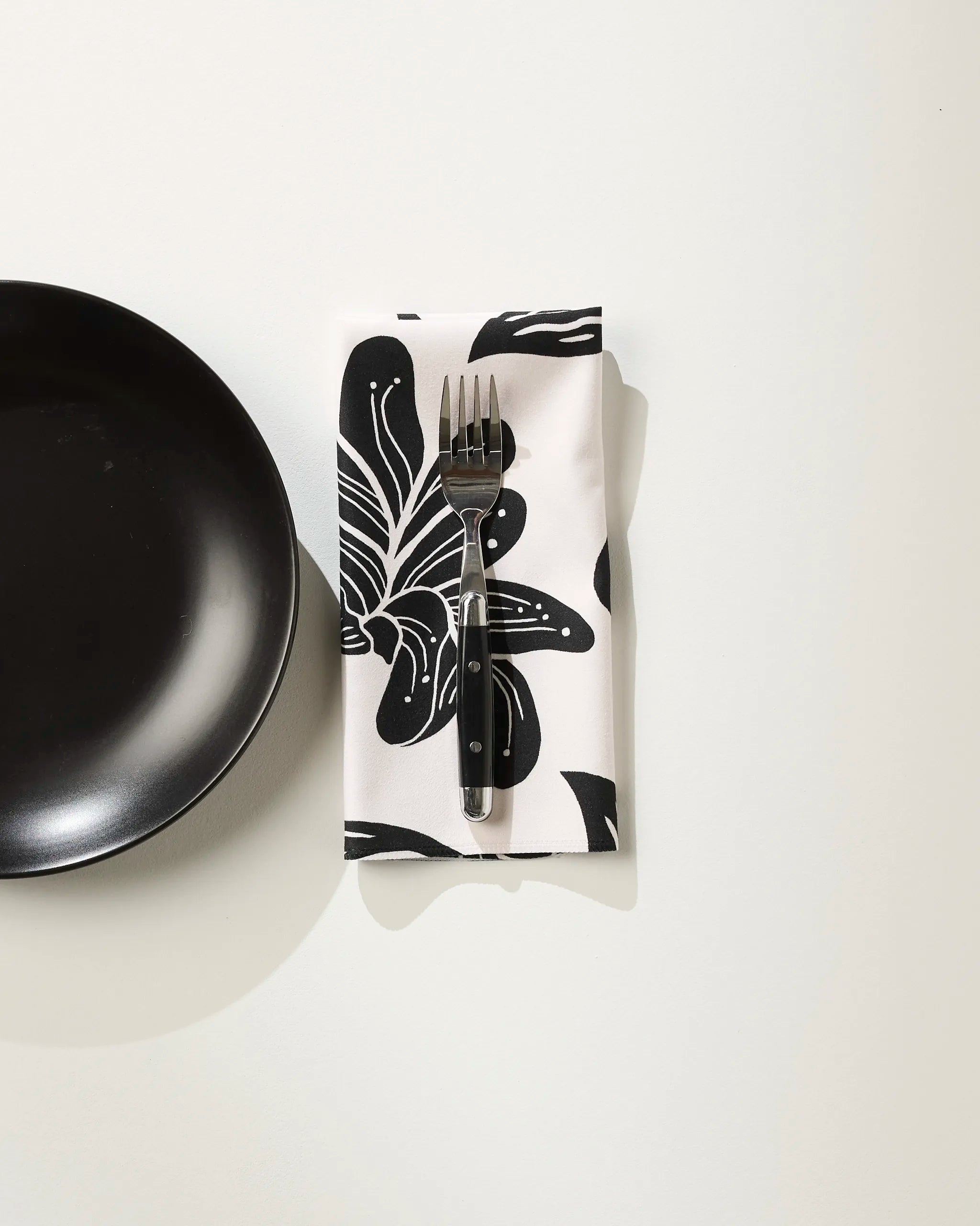 A black plate rests on a white surface beside a fork set on a folded Geometry napkin in the Giant Lush Floral pattern.