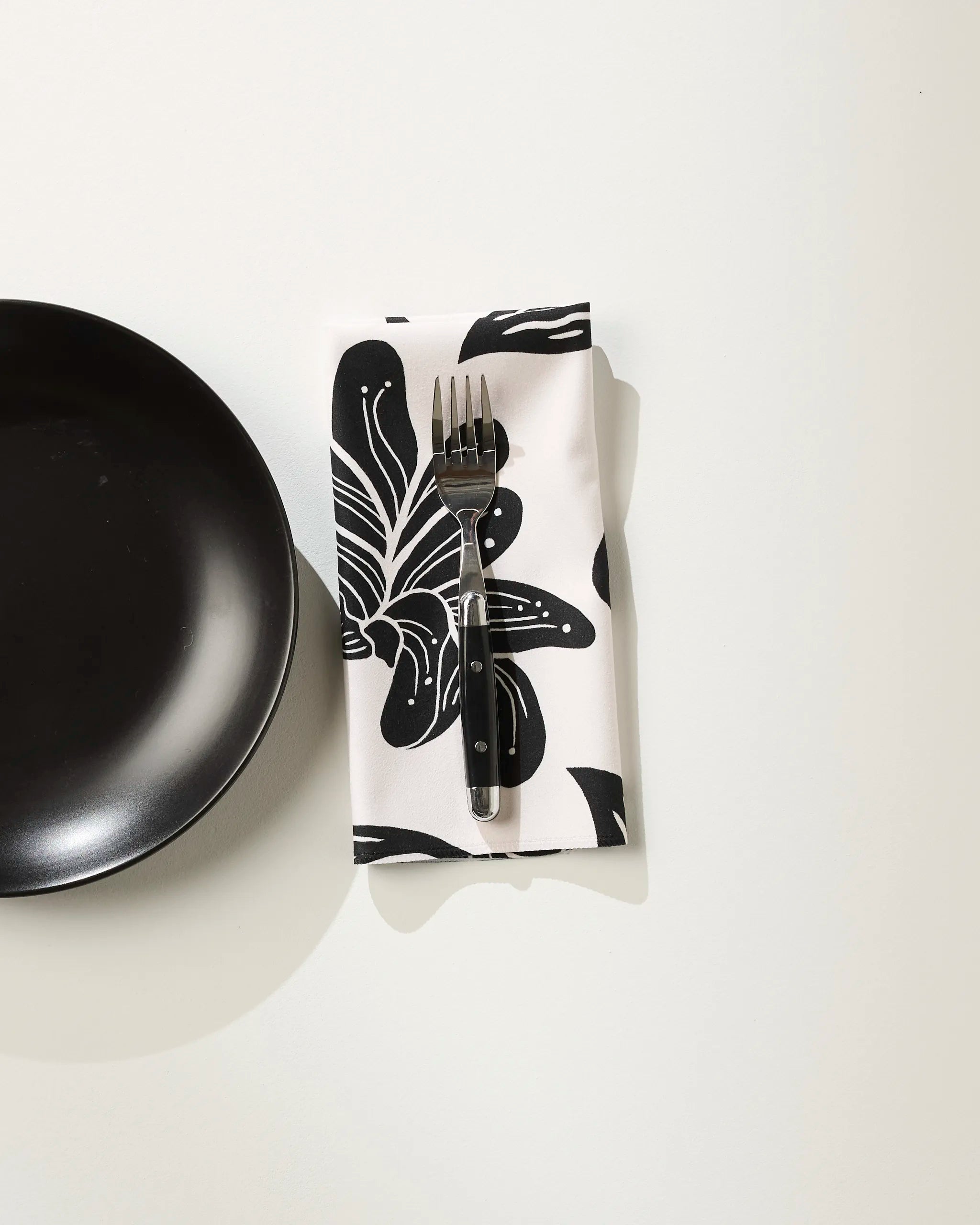 A black plate rests beside a Geometry Giant Lush Floral dinner napkin, topped with a silver fork, all neatly arranged on a white surface.
