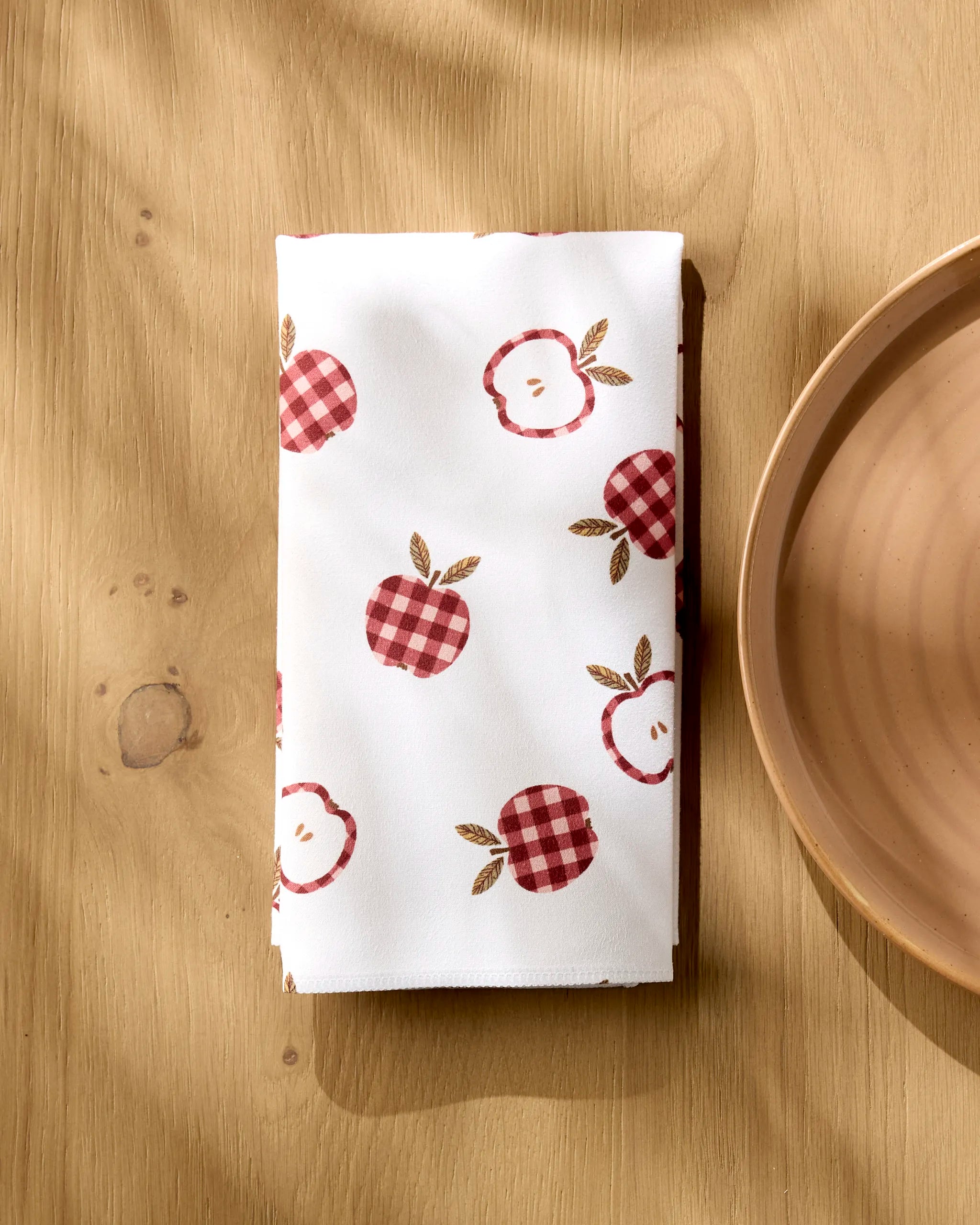 The Gingham Apples reusable white cloth napkin by Teri the Butter, featuring red apple and plaid apple designs, rests on a light wooden table beside a beige ceramic plate in gentle sunlight.