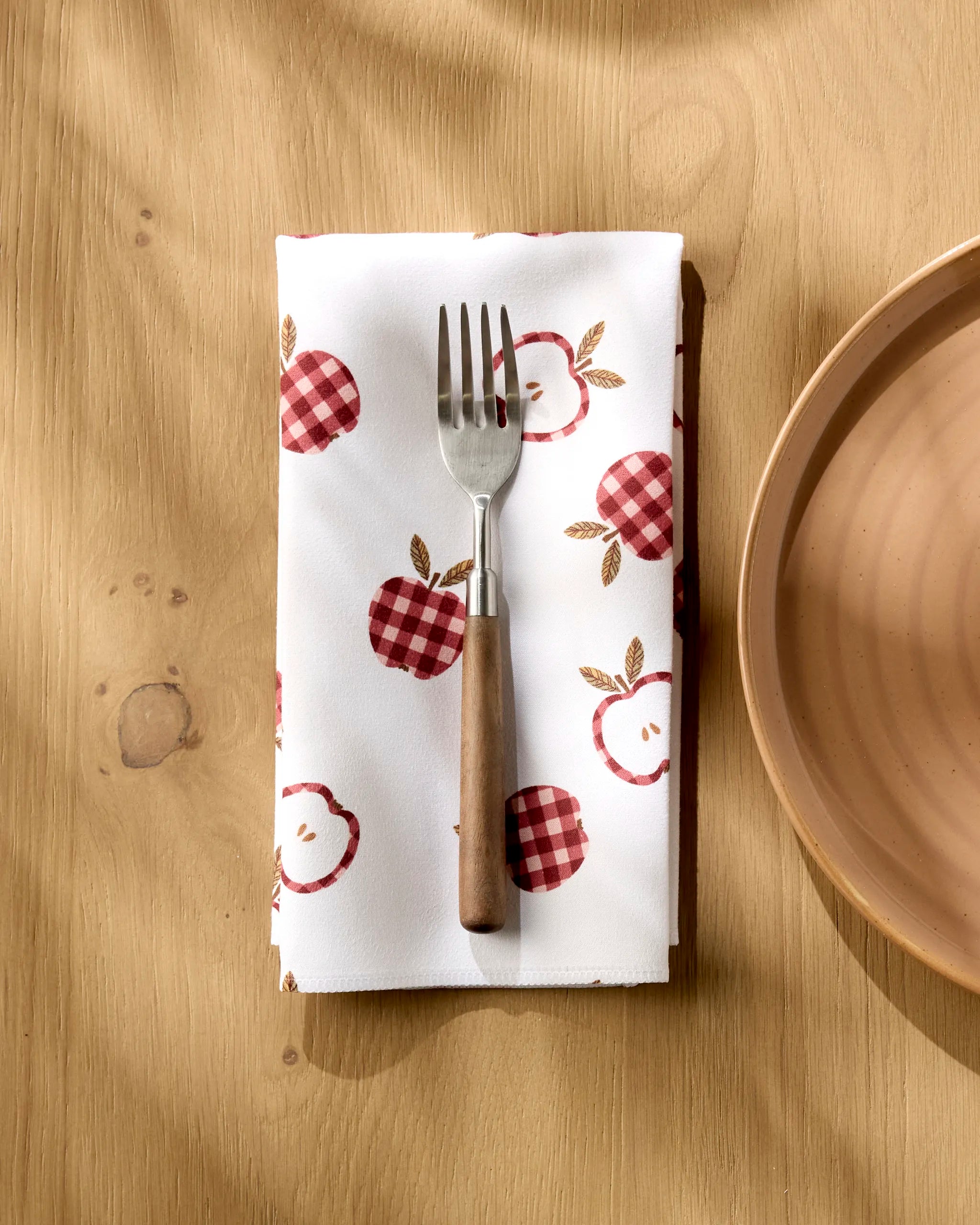 A fork with a wooden handle rests on Teri the Butter’s “Gingham Apples” reusable cloth napkin, featuring red and pink apples, beside a light brown ceramic plate on a wooden table.