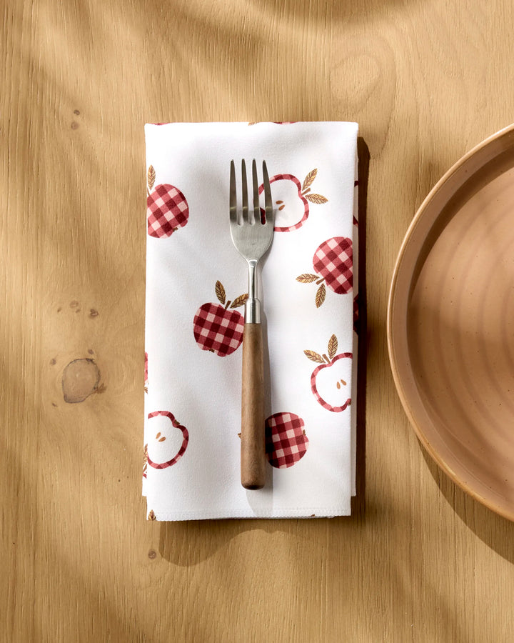 A fork with a wooden handle rests on Teri the Butter’s “Gingham Apples” reusable cloth napkin, featuring red and pink apples, beside a light brown ceramic plate on a wooden table.