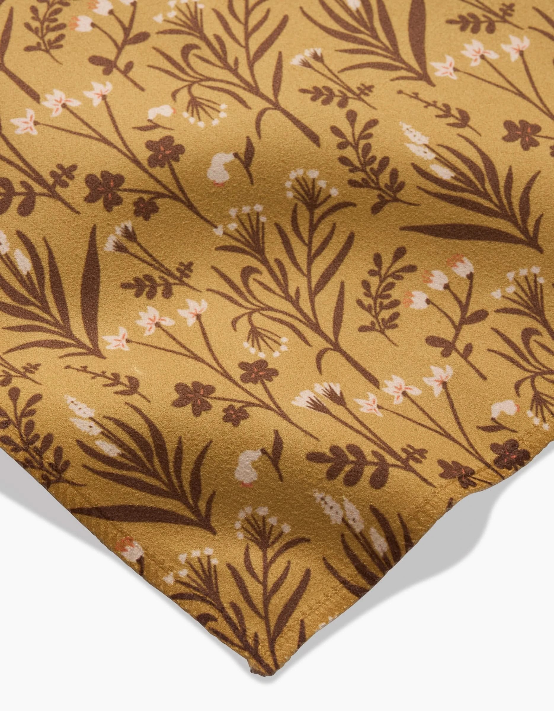 A close-up of the Golden Fall Flowers napkin by Danelys Sidron, showcasing its mustard yellow botanical pattern with brown and cream flowers and leaves against a white background.