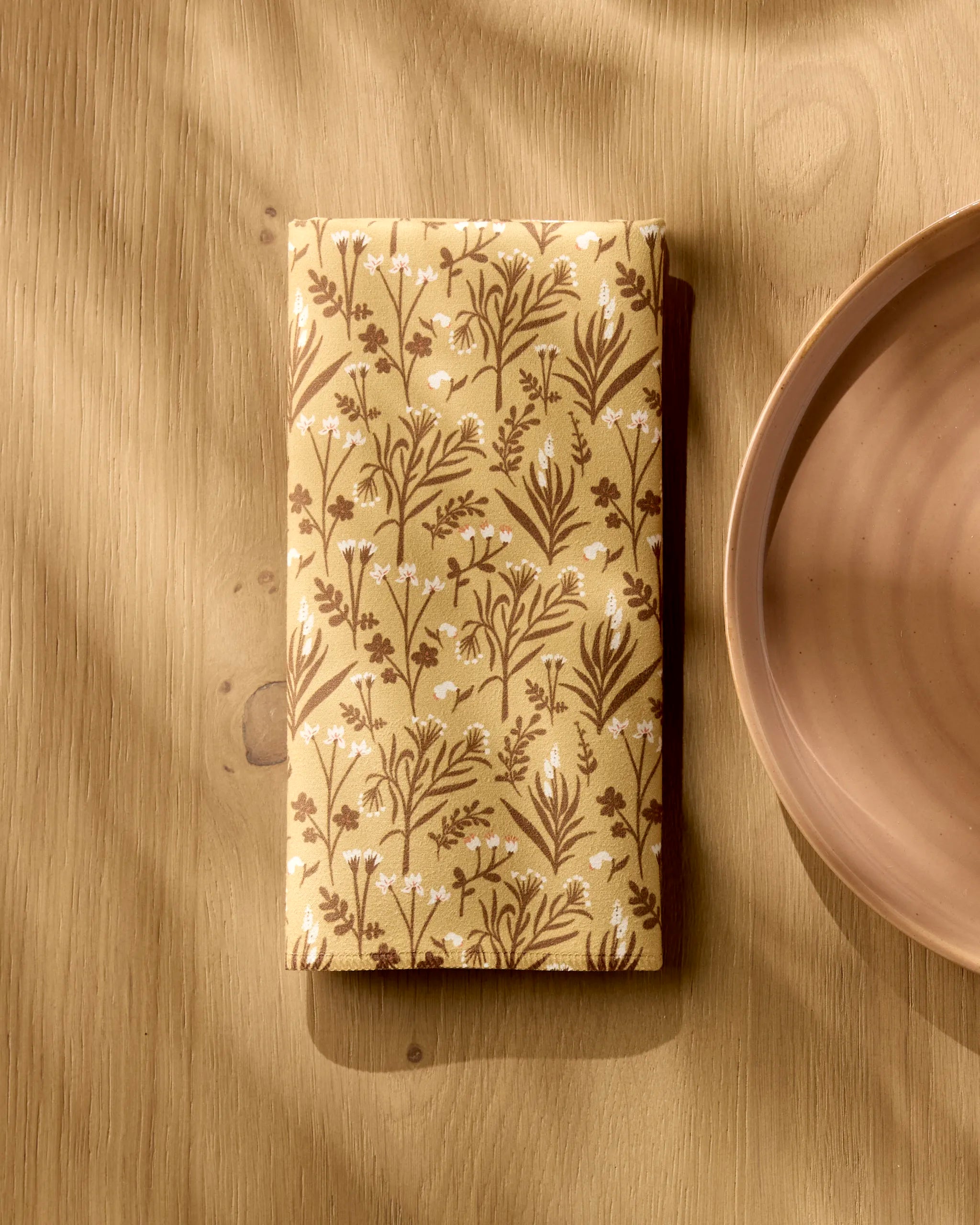 A folded yellow Golden Fall Flowers napkin by Danelys Sidron, featuring a brown and white floral pattern, rests on a light wooden table beside a round ceramic plate as sunlight softly highlights the Dinner Napkin Set.