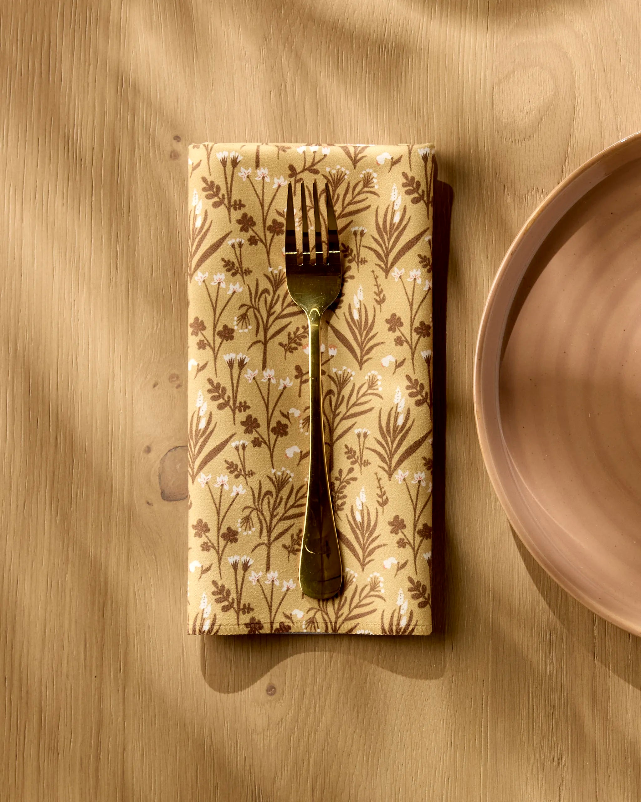 A metal fork rests on a Golden Fall Flowers napkin by Danelys Sidron, next to a plain light brown ceramic plate on a wooden table in warm sunlight.