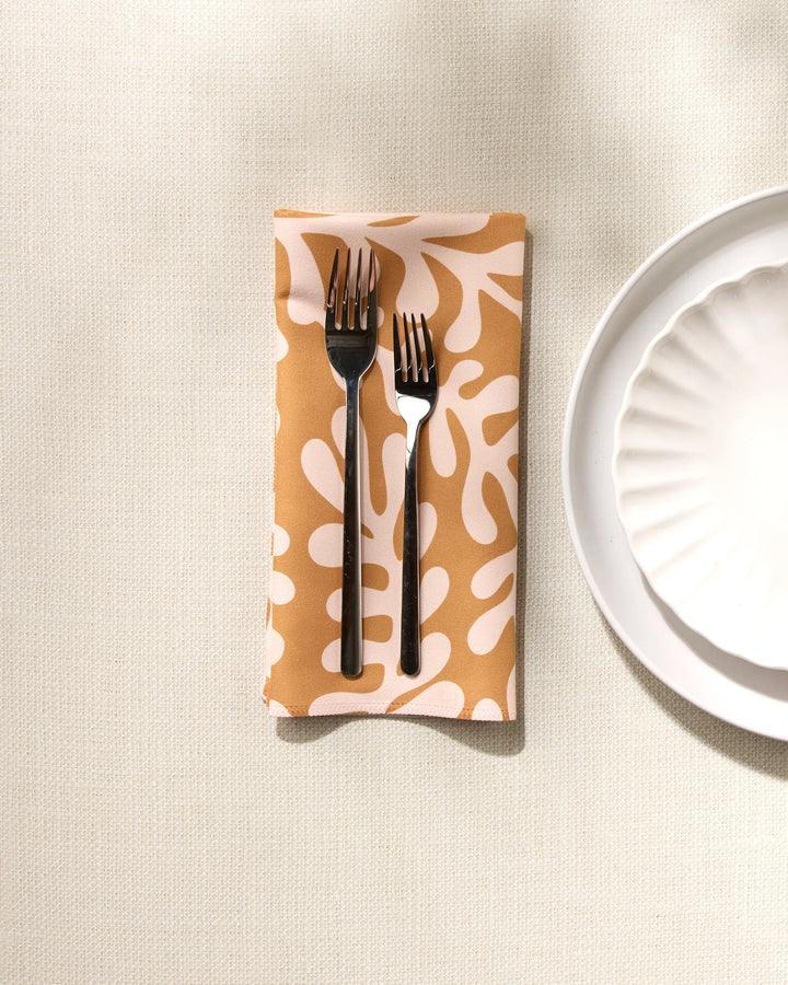 Two forks rest side by side on a folded GEOMETRY Golden Fall reusable dinner napkin with a beige and white abstract pattern, next to a scalloped white plate on a light textured surface.