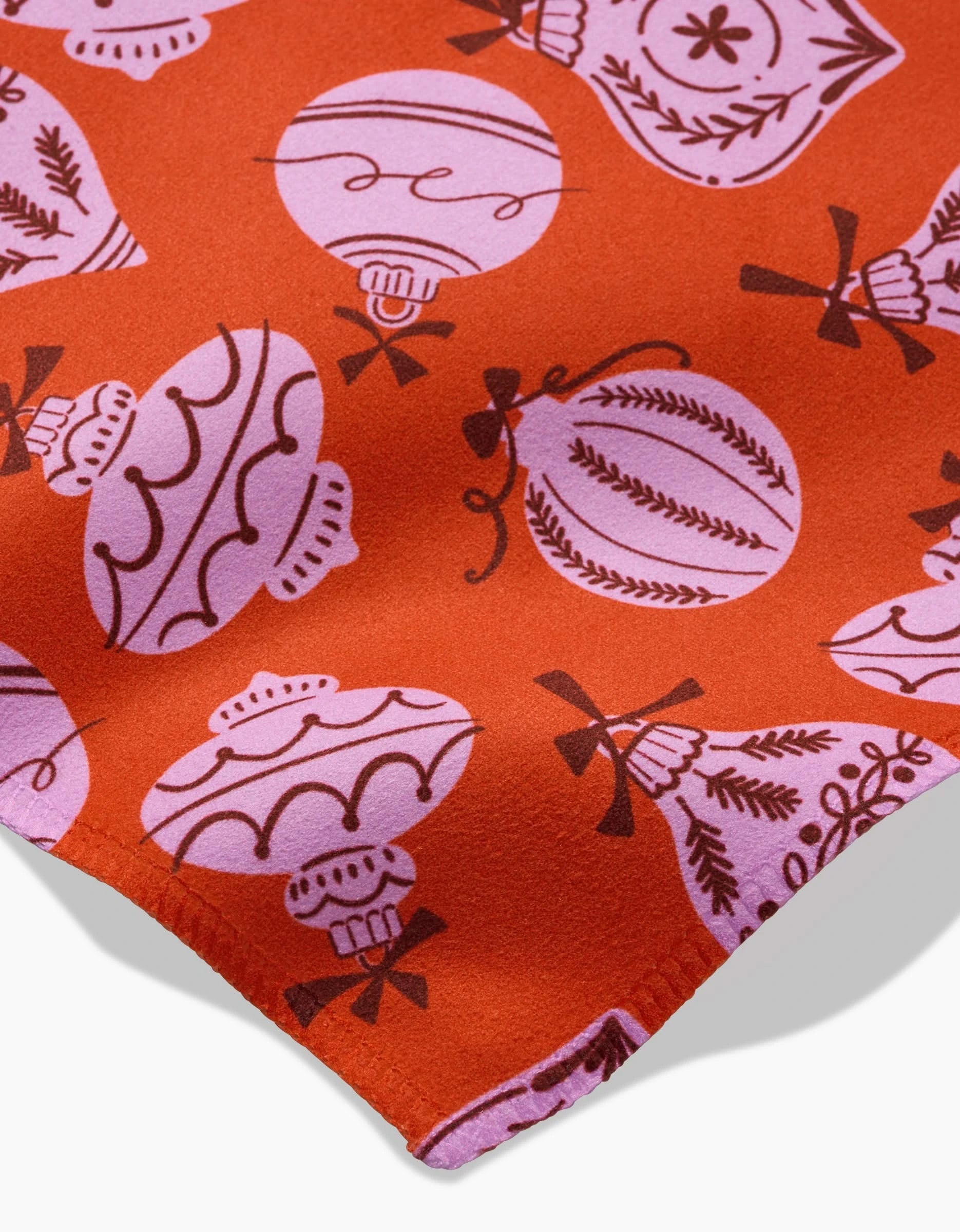 A close-up of Grans Ornaments fabric by Charlie Rowan Designs shows orange material with pink and purple Christmas ornaments—ideal for festive reusable cloth napkins. The edge appears in the corner, highlighting its decorative design.