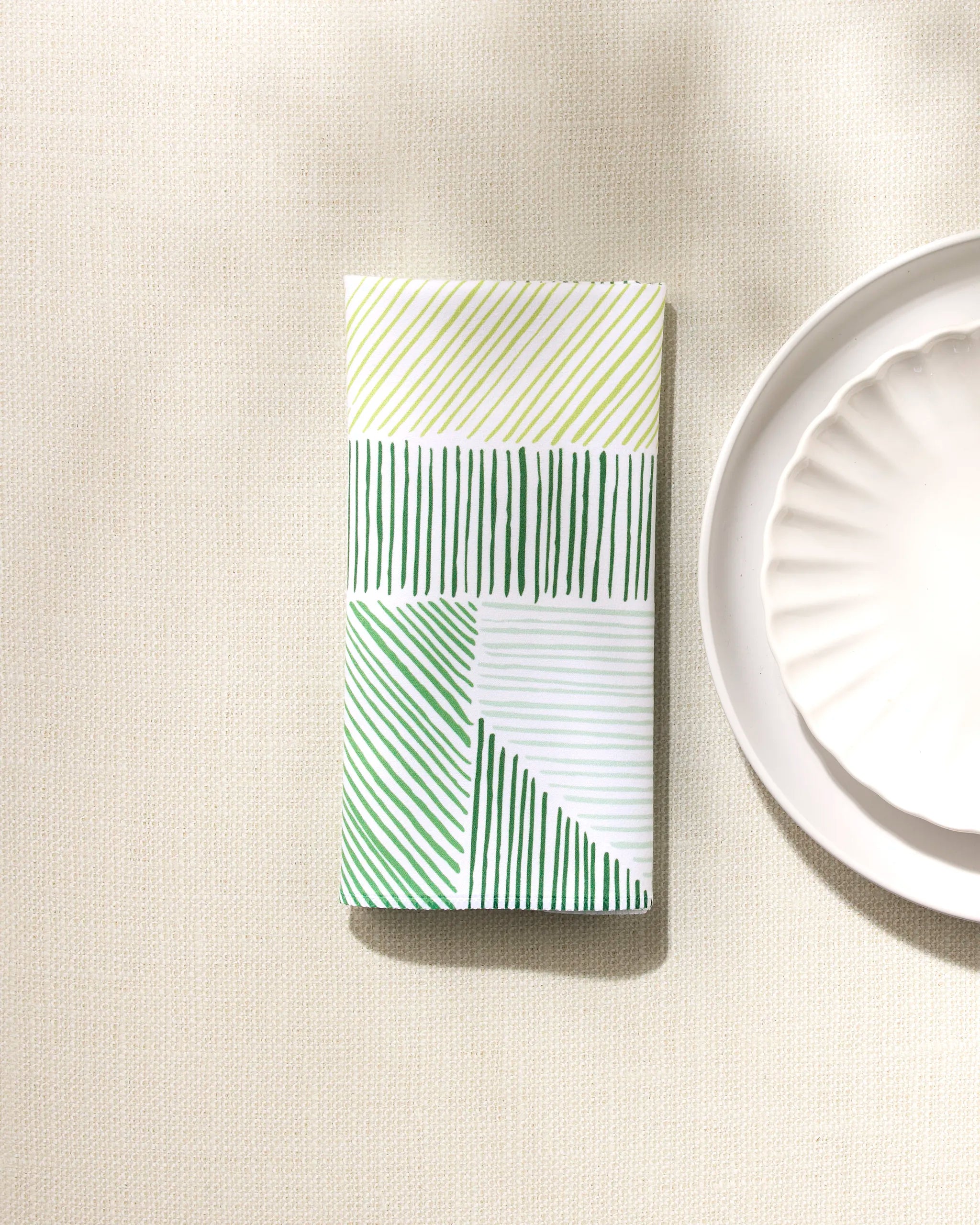 A folded GEOMETRY Green Kites eco-friendly napkin with green and yellow diagonal and vertical lines sits beside a white scalloped-edged plate on a light textured surface.