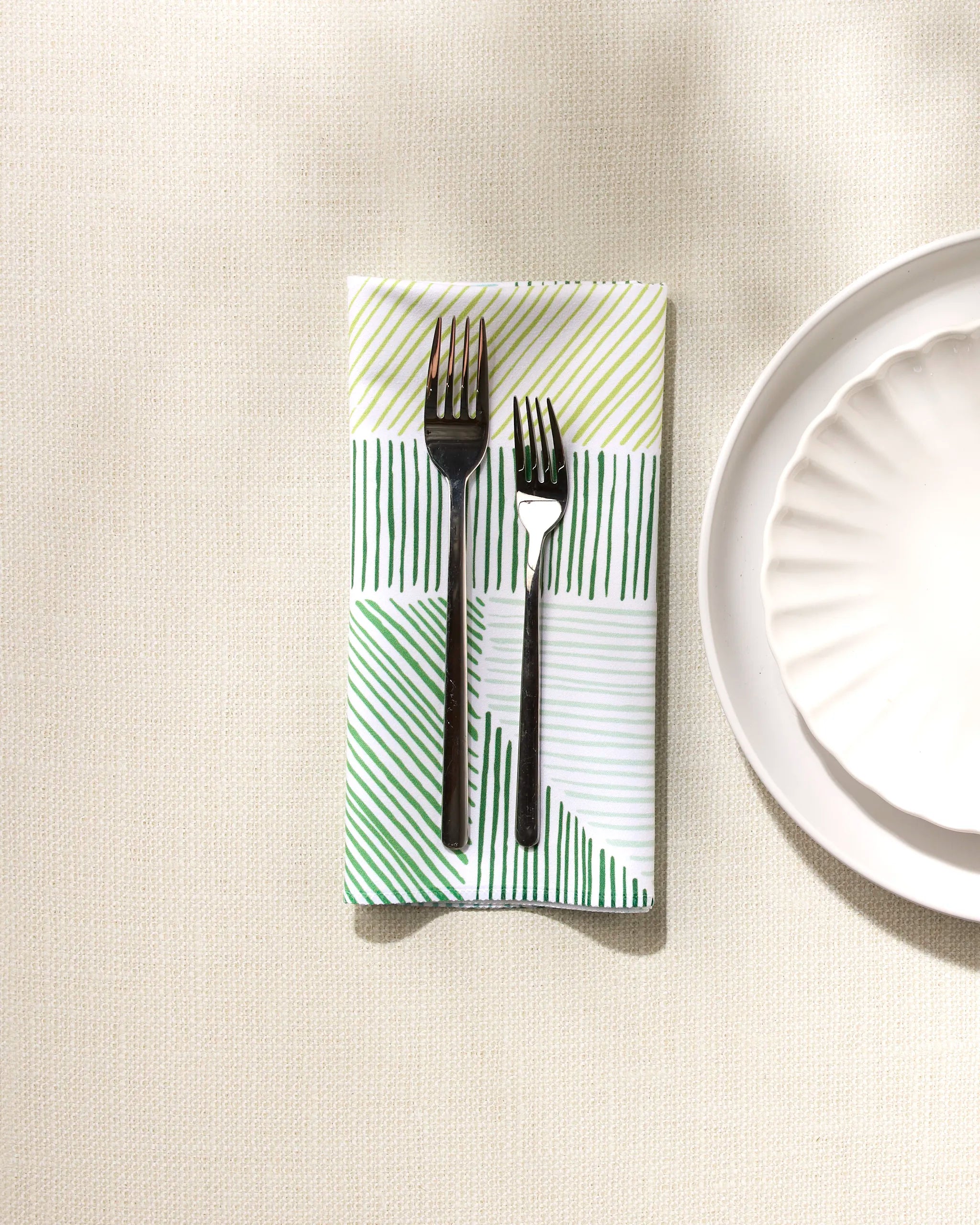 A table setting featuring GEOMETRY's Green Kites eco-friendly napkin with green diagonal stripes, folded to hold a knife and fork atop a white plate, all placed on a textured cream-colored surface.