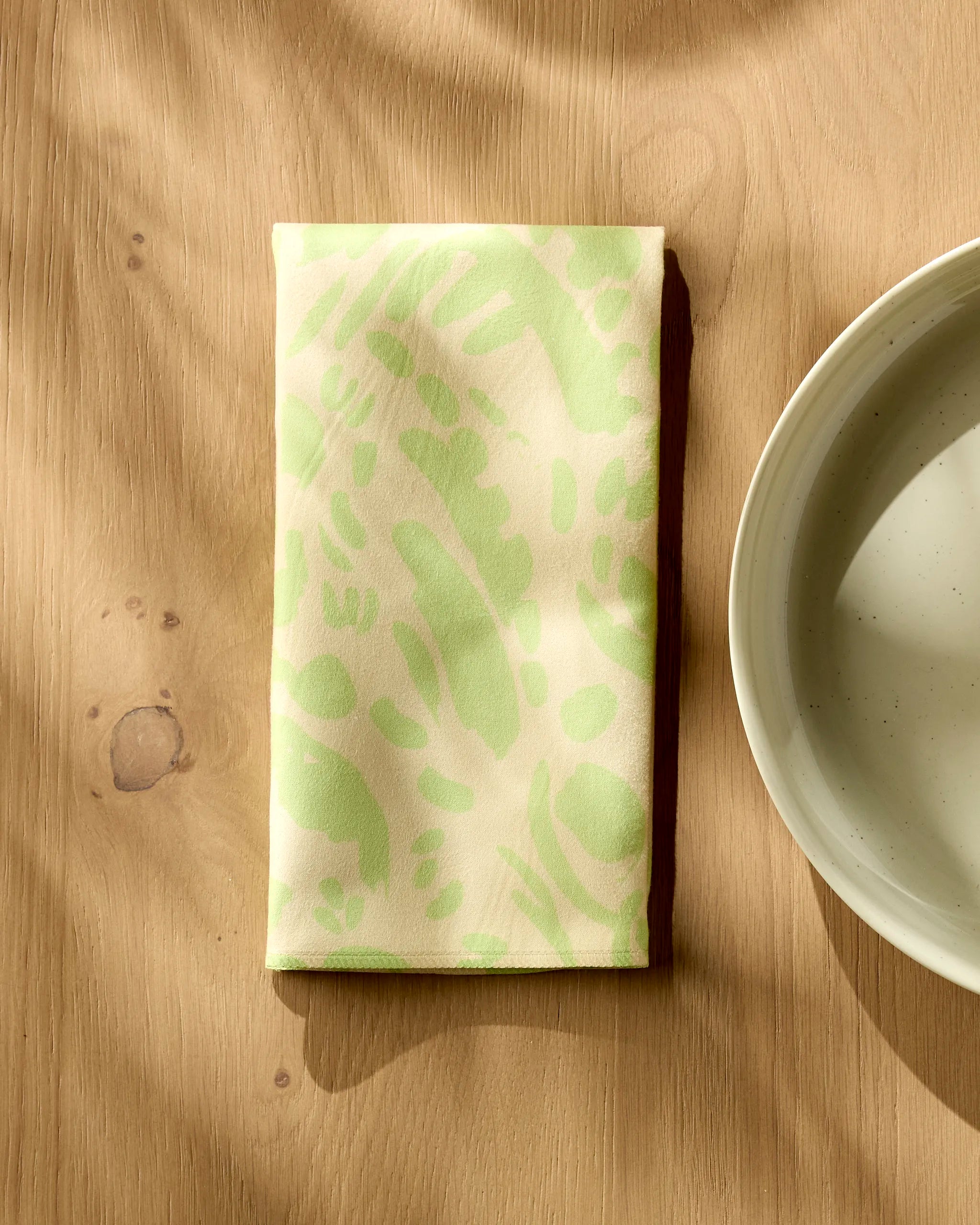 A folded Sareka Unique "Green Spring" recycled napkin with a pale green abstract pattern rests on a light wooden table beside a white ceramic bowl, soft shadows falling across the scene.
