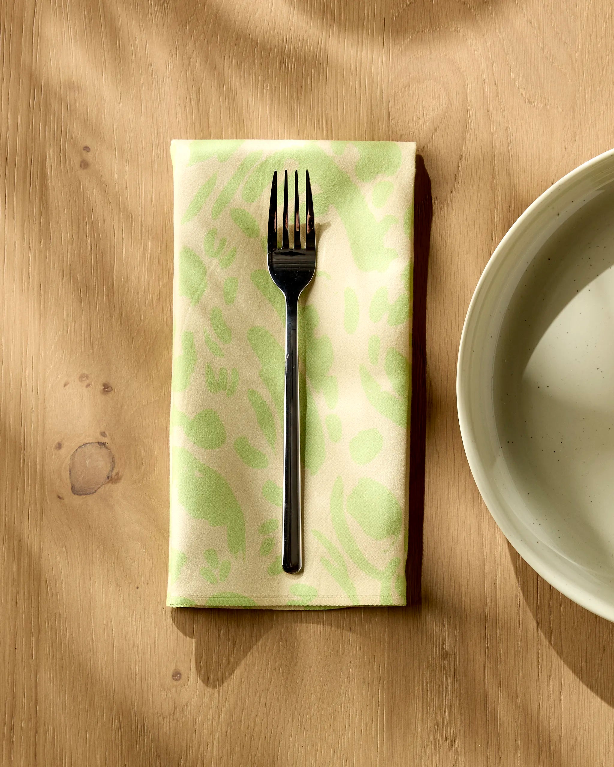 A Sareka Unique Green Spring metal fork sits atop a folded reusable dinner napkin on a wooden table beside a white ceramic bowl, with soft sunlight casting gentle shadows.