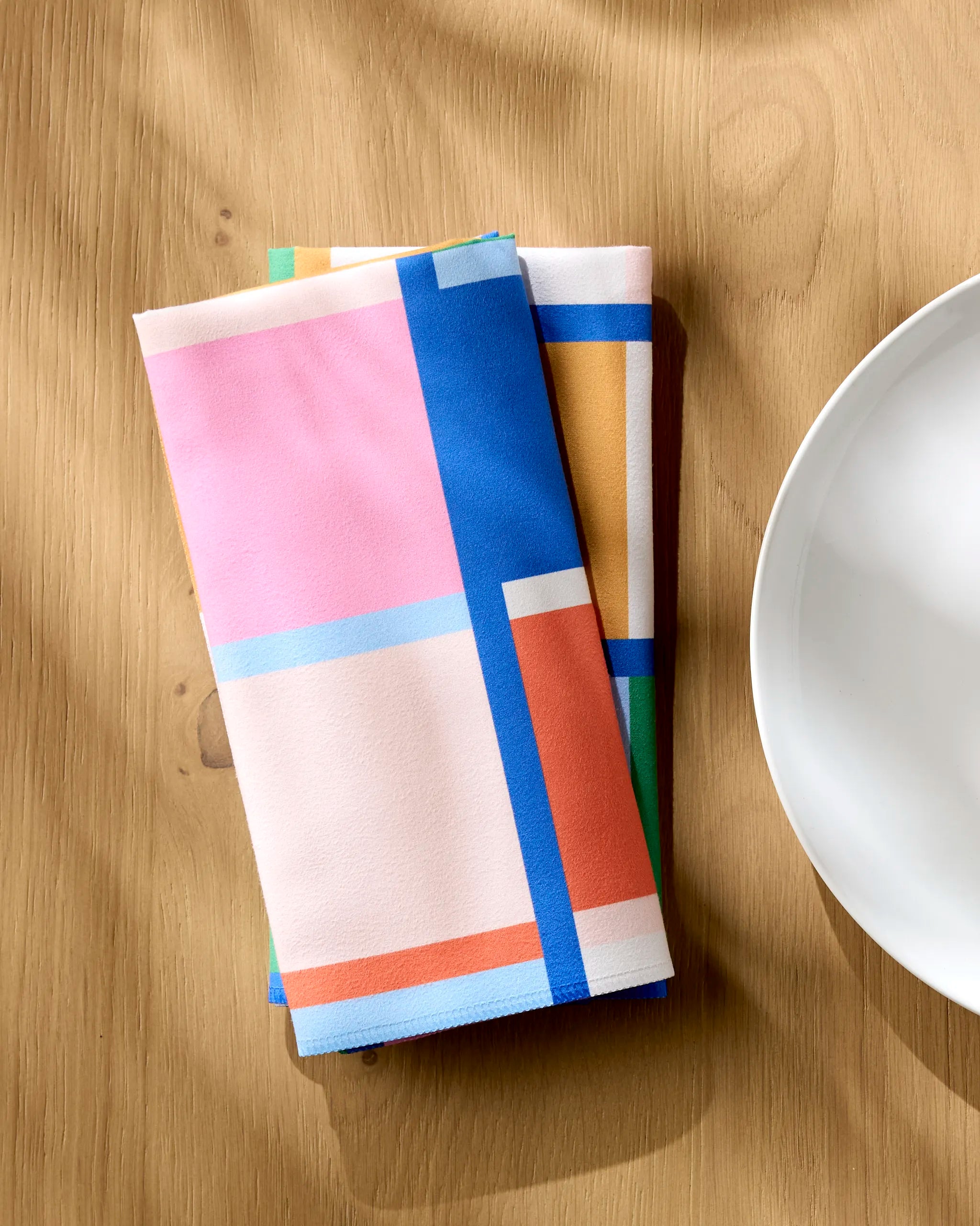 Two folded Grid napkins by Jennifer Bouron, featuring a colorful geometric pattern in pink, blue, orange, and yellow, rest on a light wooden table beside a white plate. Sunlight highlights this stylish Dinner Napkin Set.