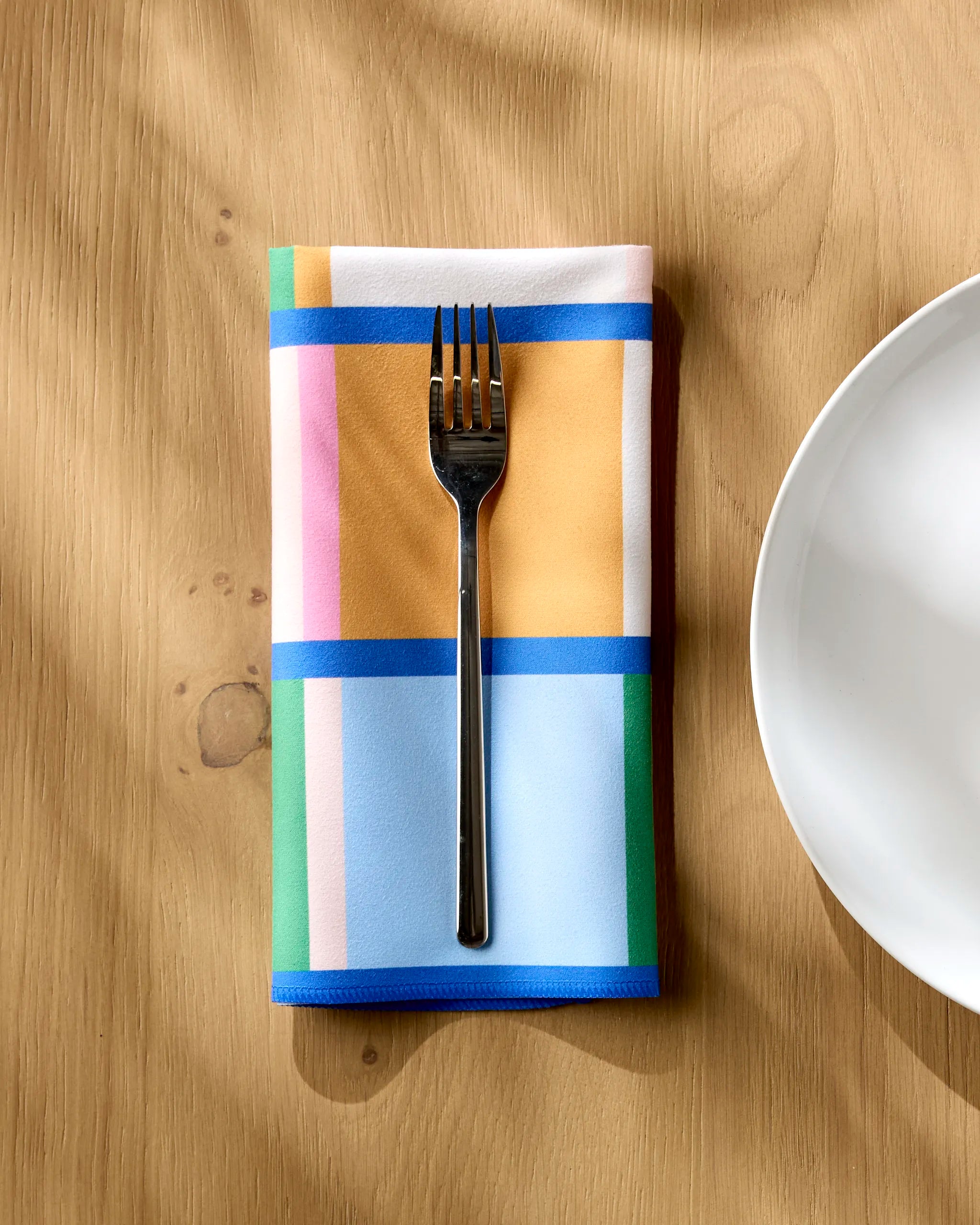 A fork rests on an absorbent "Grid" napkin by Jennifer Bouron, featuring blue, pink, yellow, and green geometric patterns, next to a white plate on a wooden table.