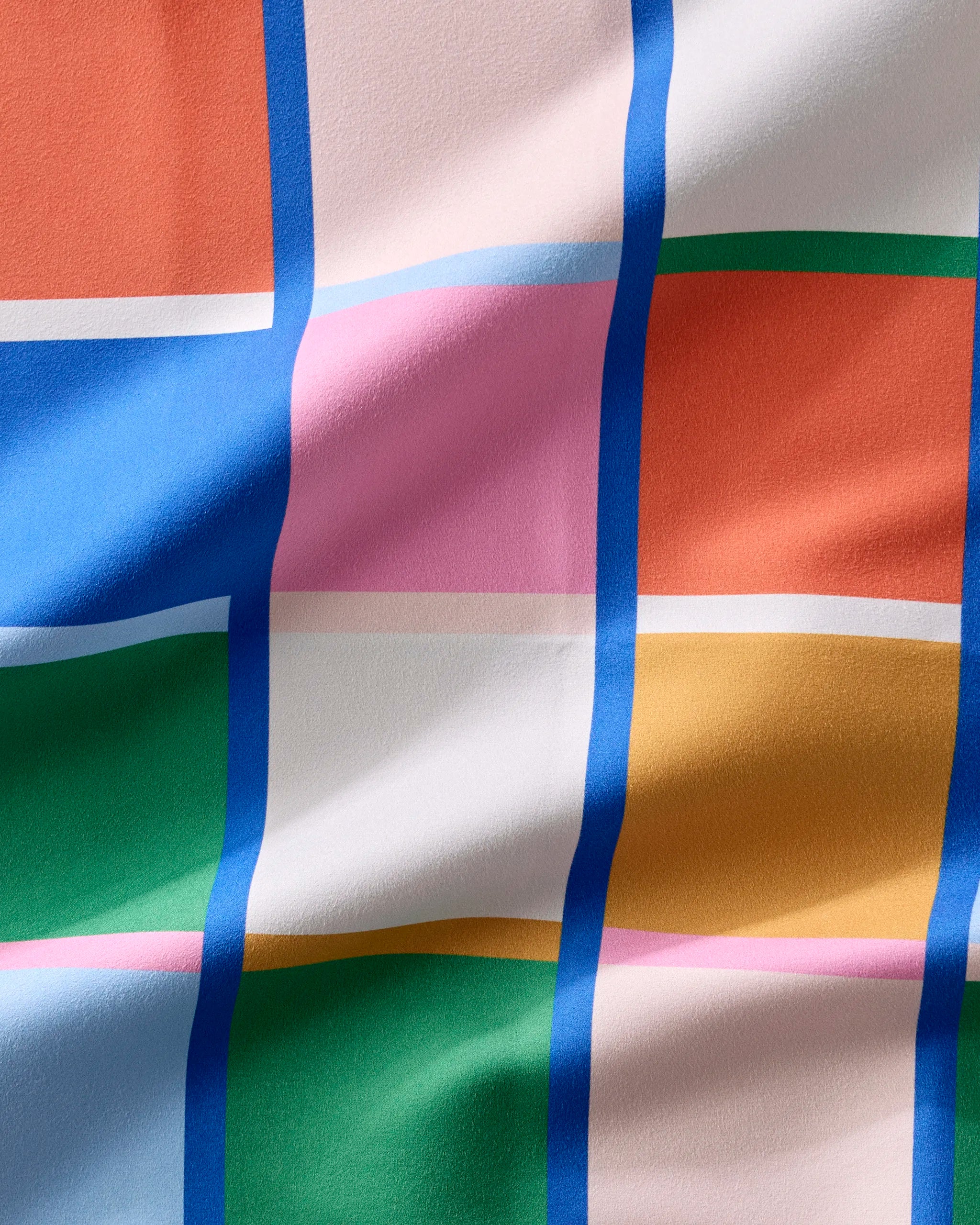 A close-up of the Grid dinner napkins by Jennifer Bouron shows absorbent recycled fabric with a colorful grid of blue, pink, orange, yellow, and green squares. Bold blue and white lines create a wavy texture across the gentle folds.