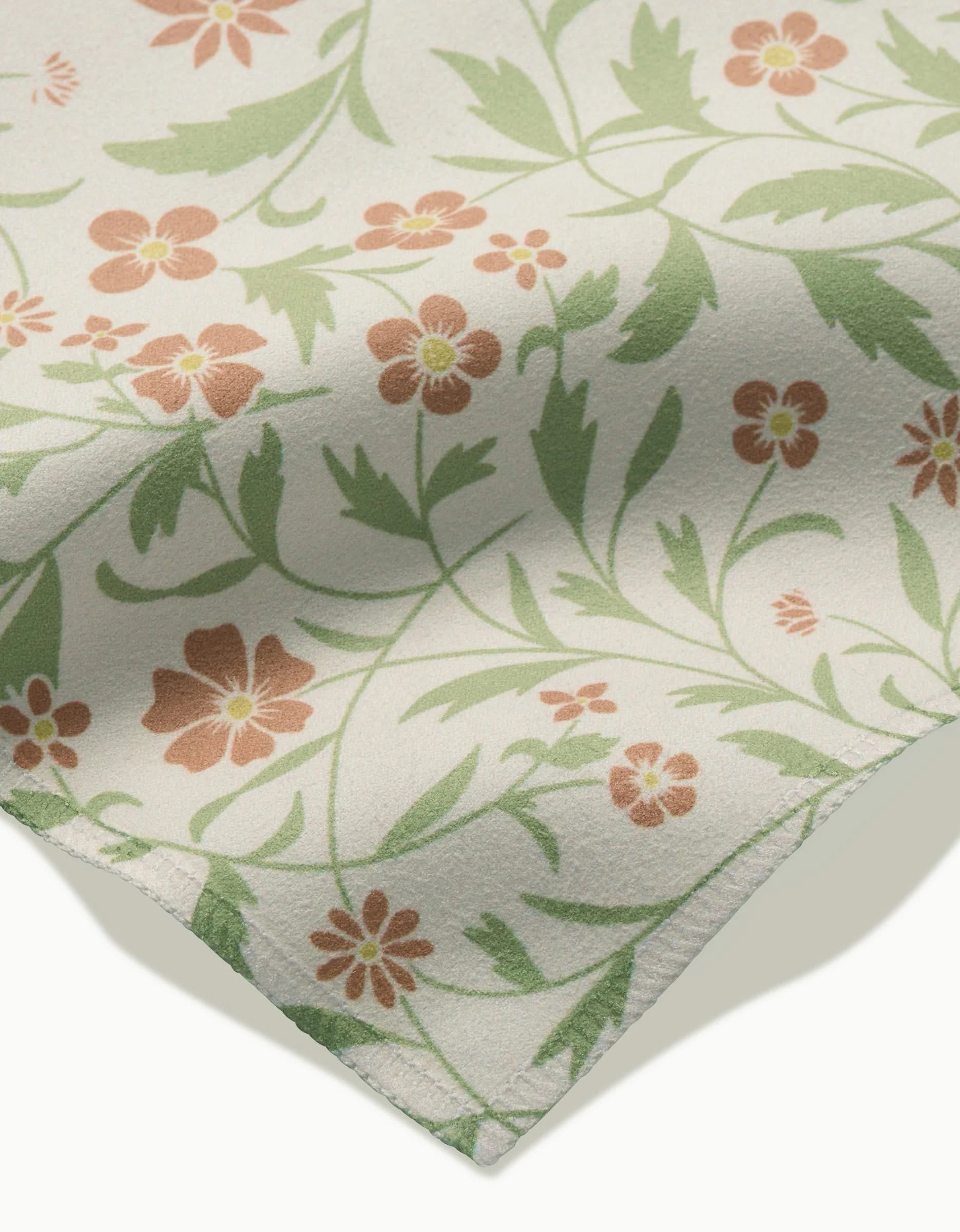 A close-up of the Hawthorns dinner napkin by Danelys Sidron, in cream Geosuede fabric, featuring a floral pattern with green leaves and orange-brown flowers, shown with a slightly folded corner.