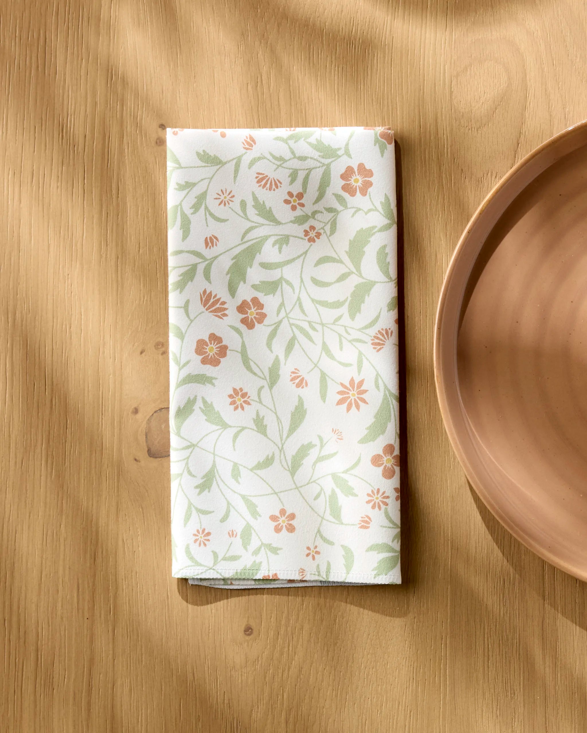 A folded white cloth napkin from Danelys Sidron's eco-friendly Hawthorns collection, featuring a green and orange floral pattern, rests on a light wooden table beside a peach ceramic plate.