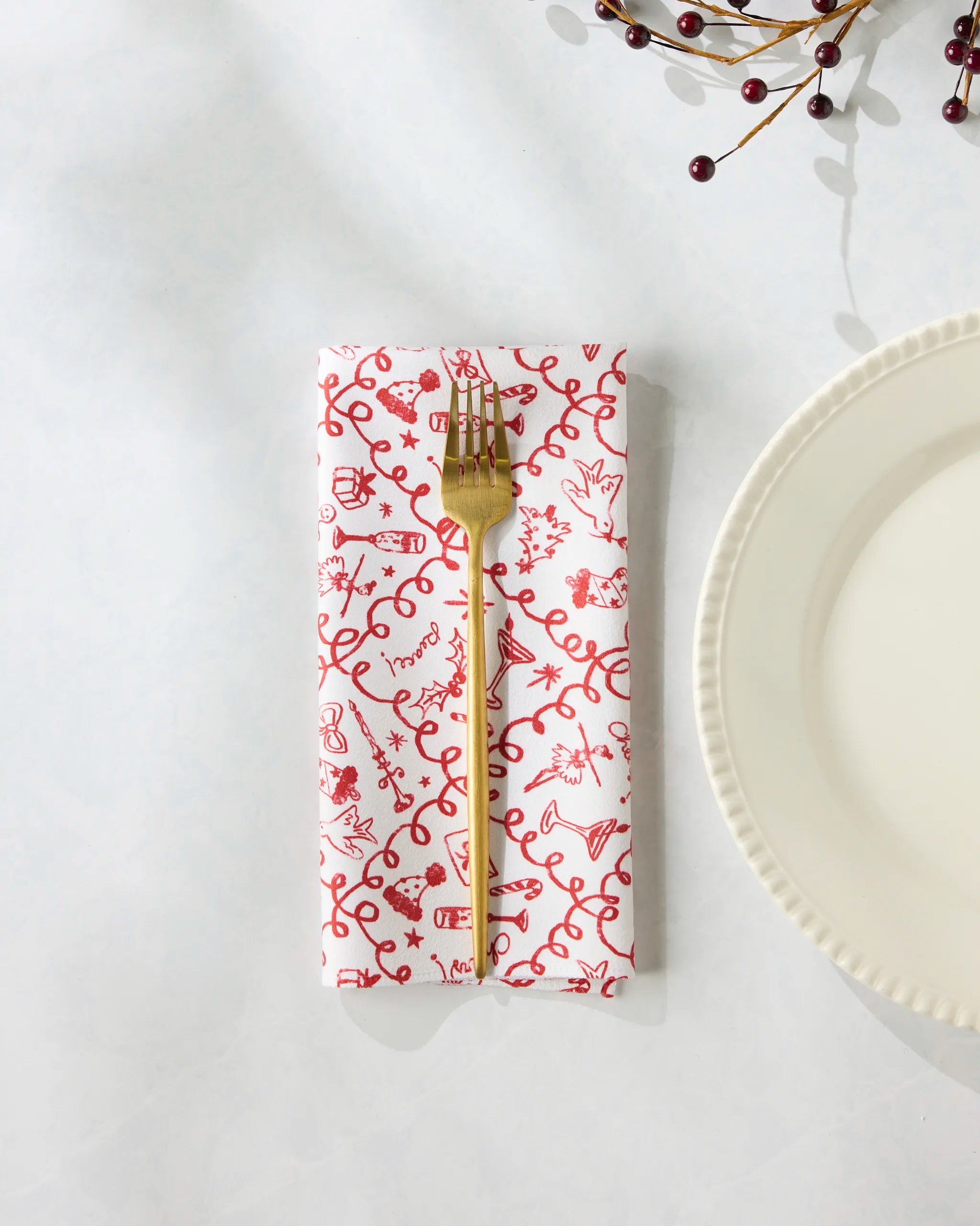 A gold fork rests on Geometry’s Holiday Doodles eco-friendly napkin with a red-and-white pattern next to a white plate, while red berries on a branch appear in the photo’s top right corner.