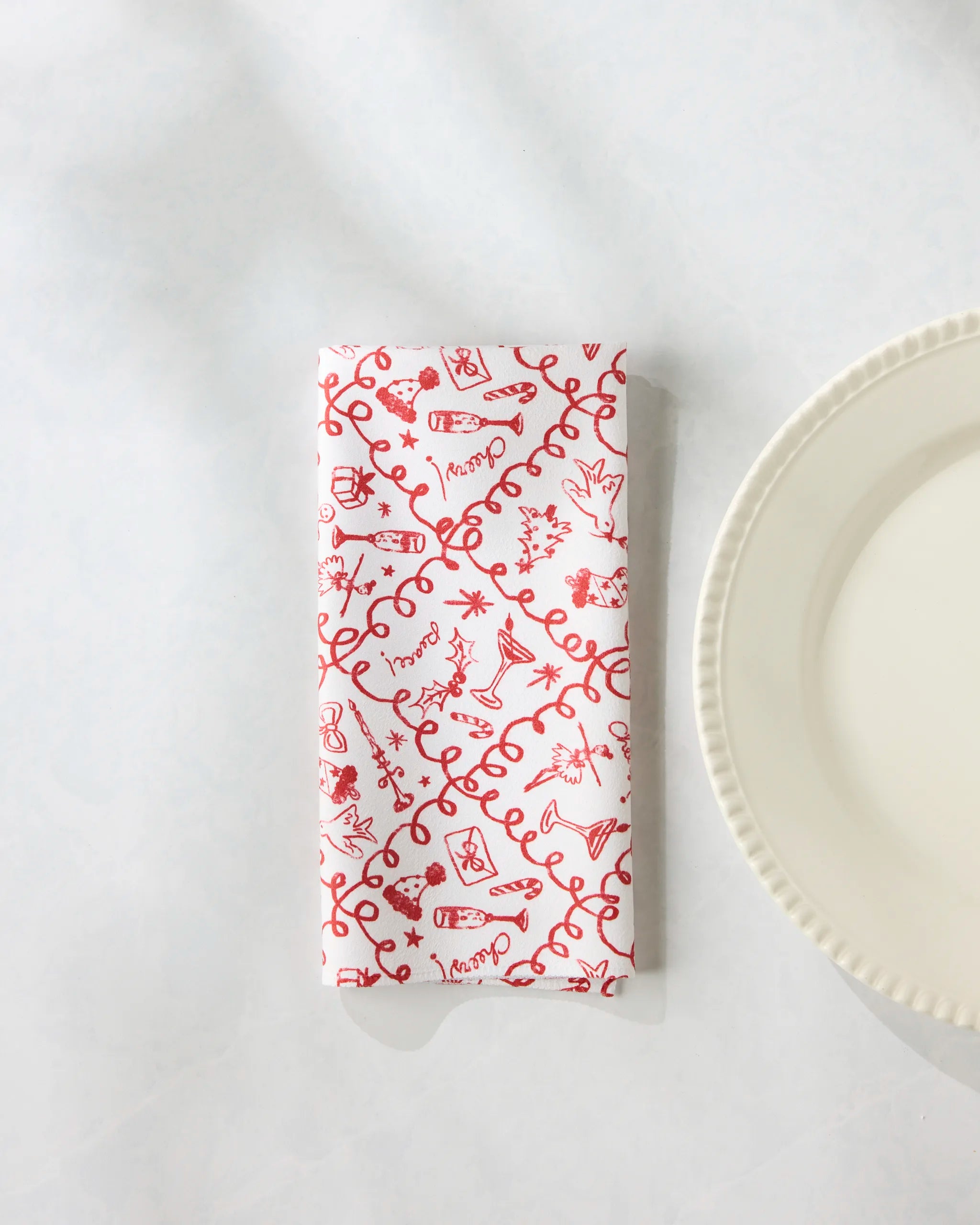 The Geometry "Holiday Doodles" eco-friendly white napkin with red decorative patterns rests on a white surface beside the edge of a round, scalloped white plate.