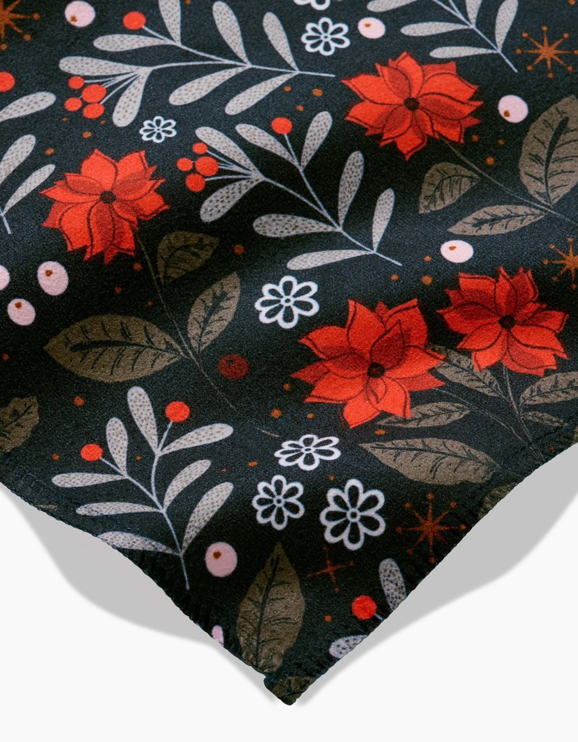 A close-up of the Holiday Floral napkin set by Charlie Rowan Designs shows dark fabric with red flowers, green leaves, white dots, small orange accents, and neatly stitched edges.