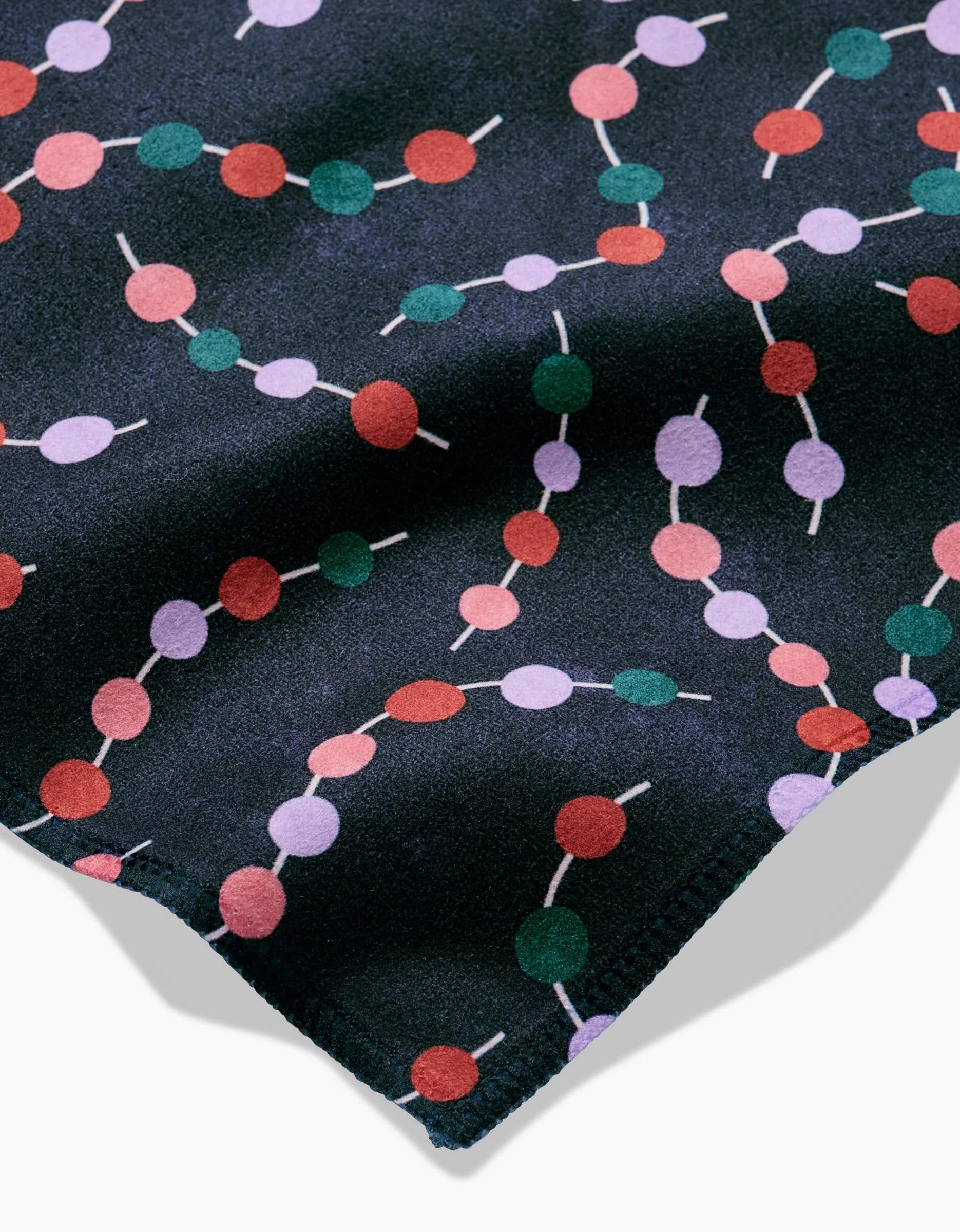 A close-up of the Claire Iglesias Holiday Garland Dinner Napkin Set in Geosuede shows a playful red, green, pink, and lavender connected circle pattern on a dark background. A folded edge reveals the soft texture of the fabric.