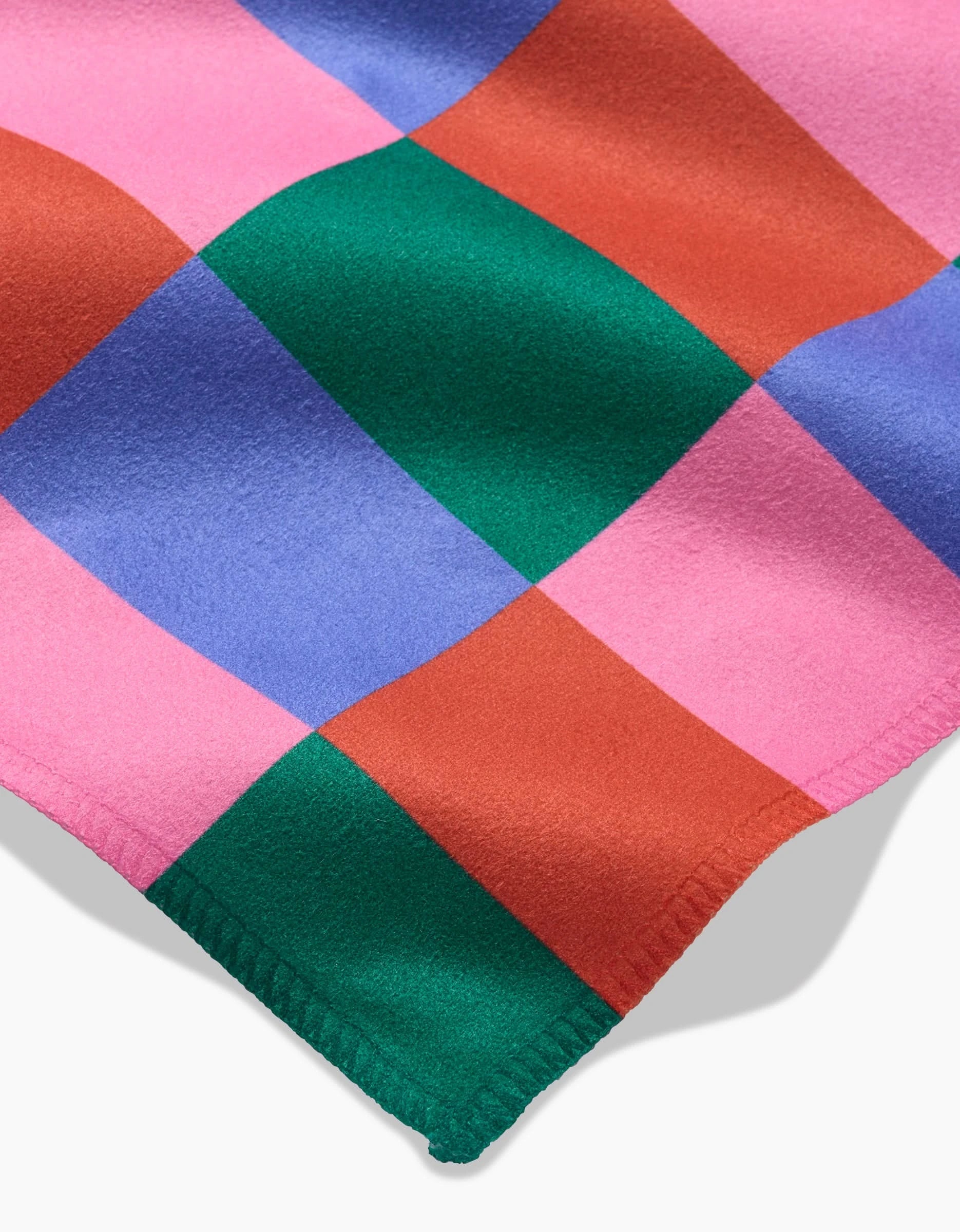 A close-up of the Laura Supnik Holiday Patchwork blanket shows its bold pink, red, green, and blue checked design. The soft fabric has stitched edging and is crafted from recycled materials for a unique holiday touch.