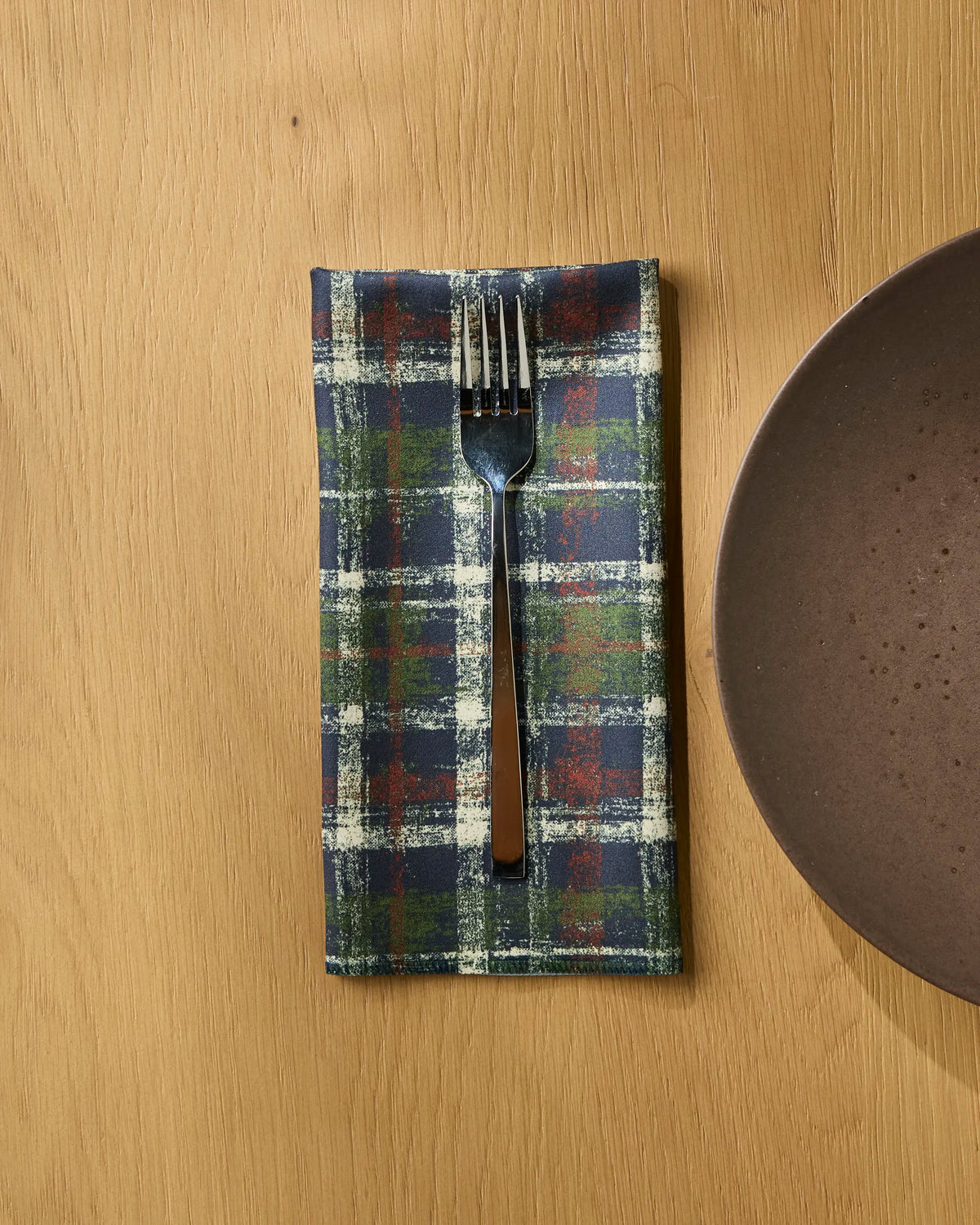 A fork rests on a folded Geometry Homestead Plaid recycled dinner napkin atop a wooden table, next to the edge of a brown ceramic plate.
