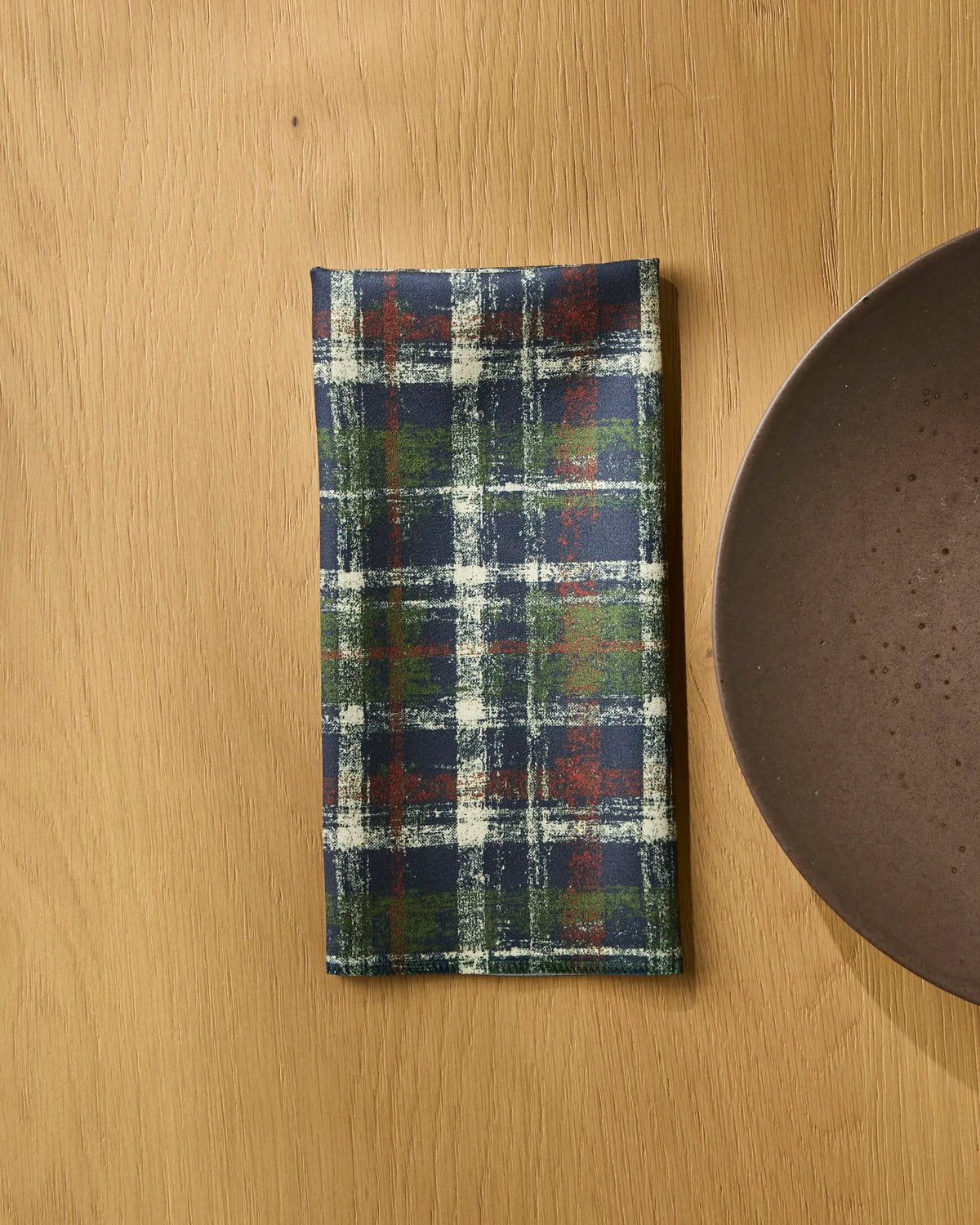 A folded Homestead Plaid dinner napkin set by Geometry, featuring blue, green, red, and white stripes, rests on a wooden table beside a brown ceramic plate. Crafted from soft Geosuede fabric, these napkins offer an elevated dining experience.
