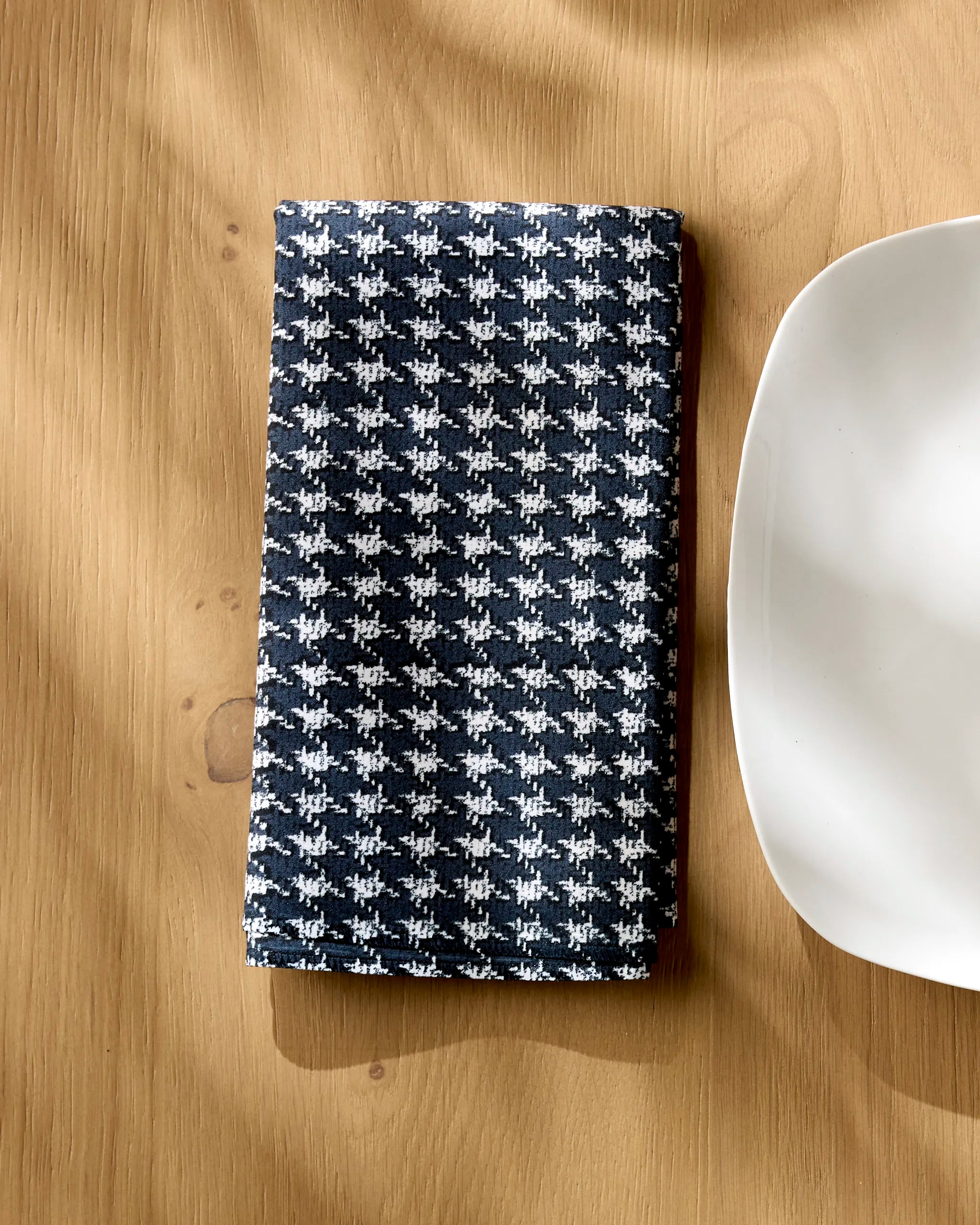 A folded Geometry Houndstooth Black napkin from our Dinner Napkin Set rests on a light wooden table beside a white square plate, with sunlight casting gentle shadows.