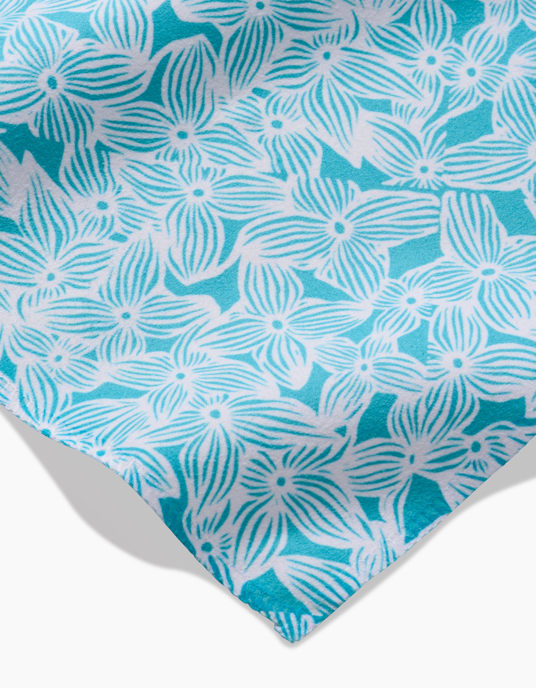 A close-up of GEOMETRY’s Hydrangea Geosuede fabric shows turquoise material with a white floral pattern, featuring overlapping flowers with striped petals and dotted centers, displayed at an angle with a visible corner.
