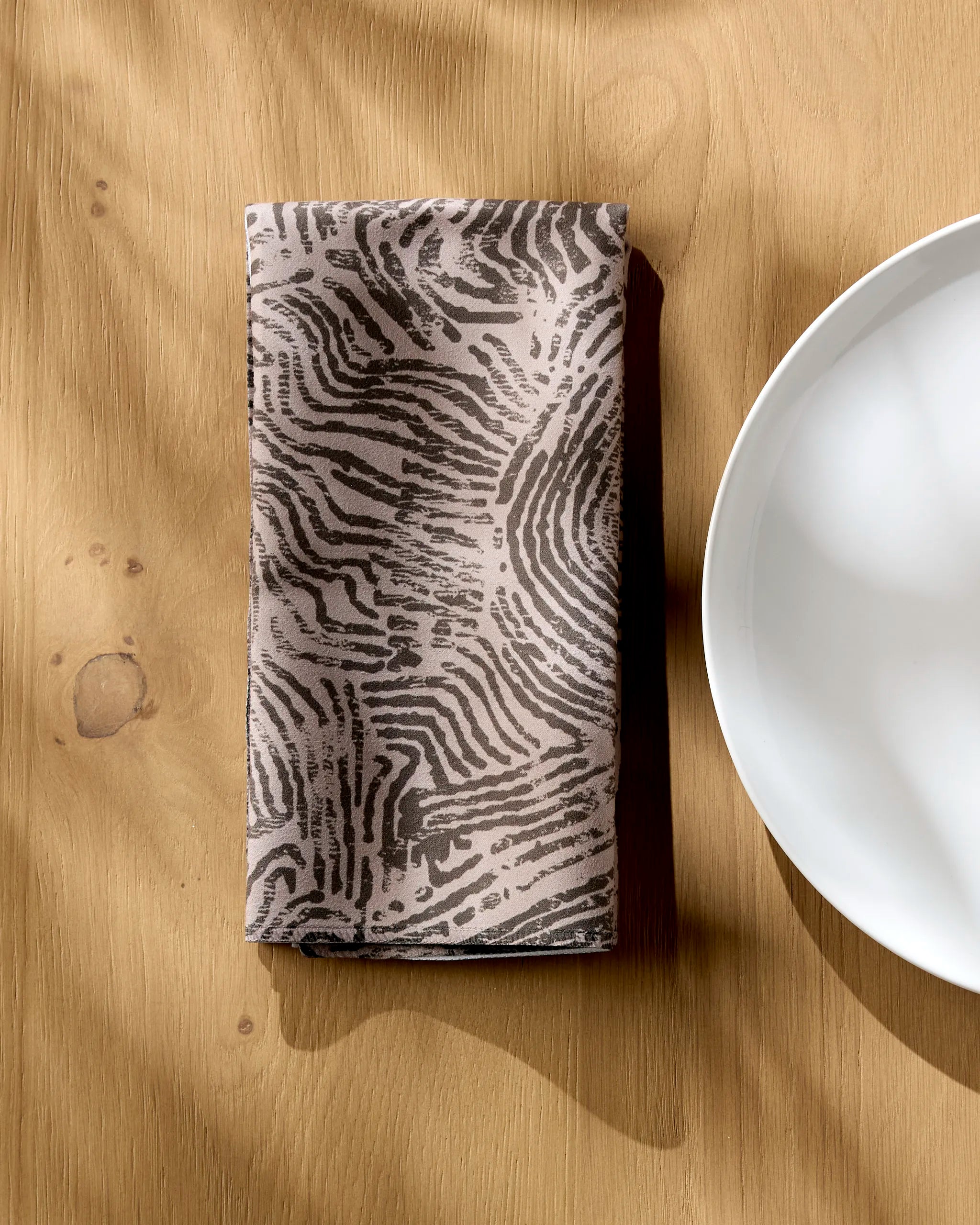 The Studio Jeannot Imprint recycled napkin, featuring a black and gray abstract design, rests folded on a wooden table beside a white plate.