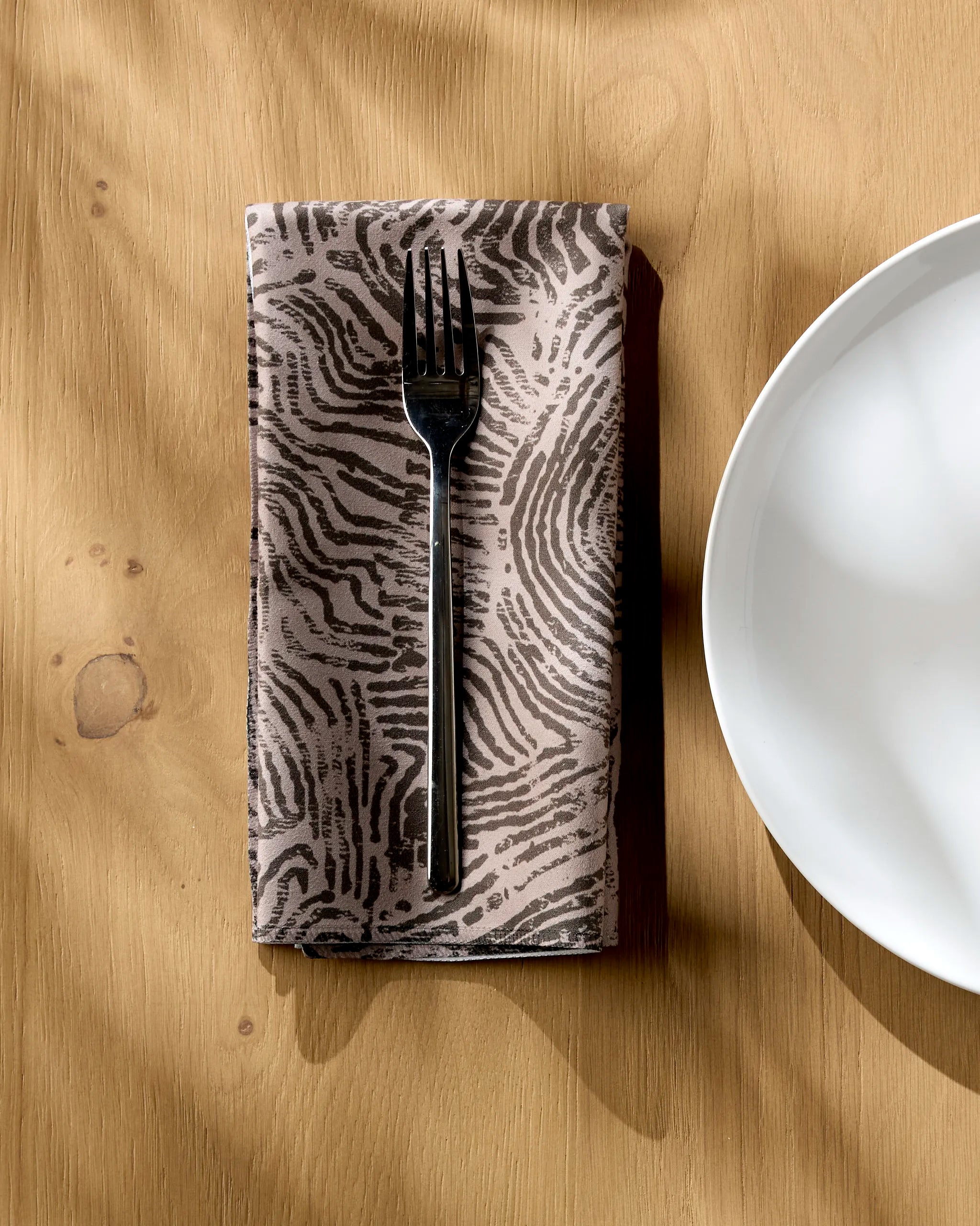 A black fork rests on Studio Jeannot's Imprint napkin—crafted from patterned Geosuede fabric with a fingerprint motif—set on a light wooden table beside a white plate, as sunlight casts soft shadows across the recycled napkin.