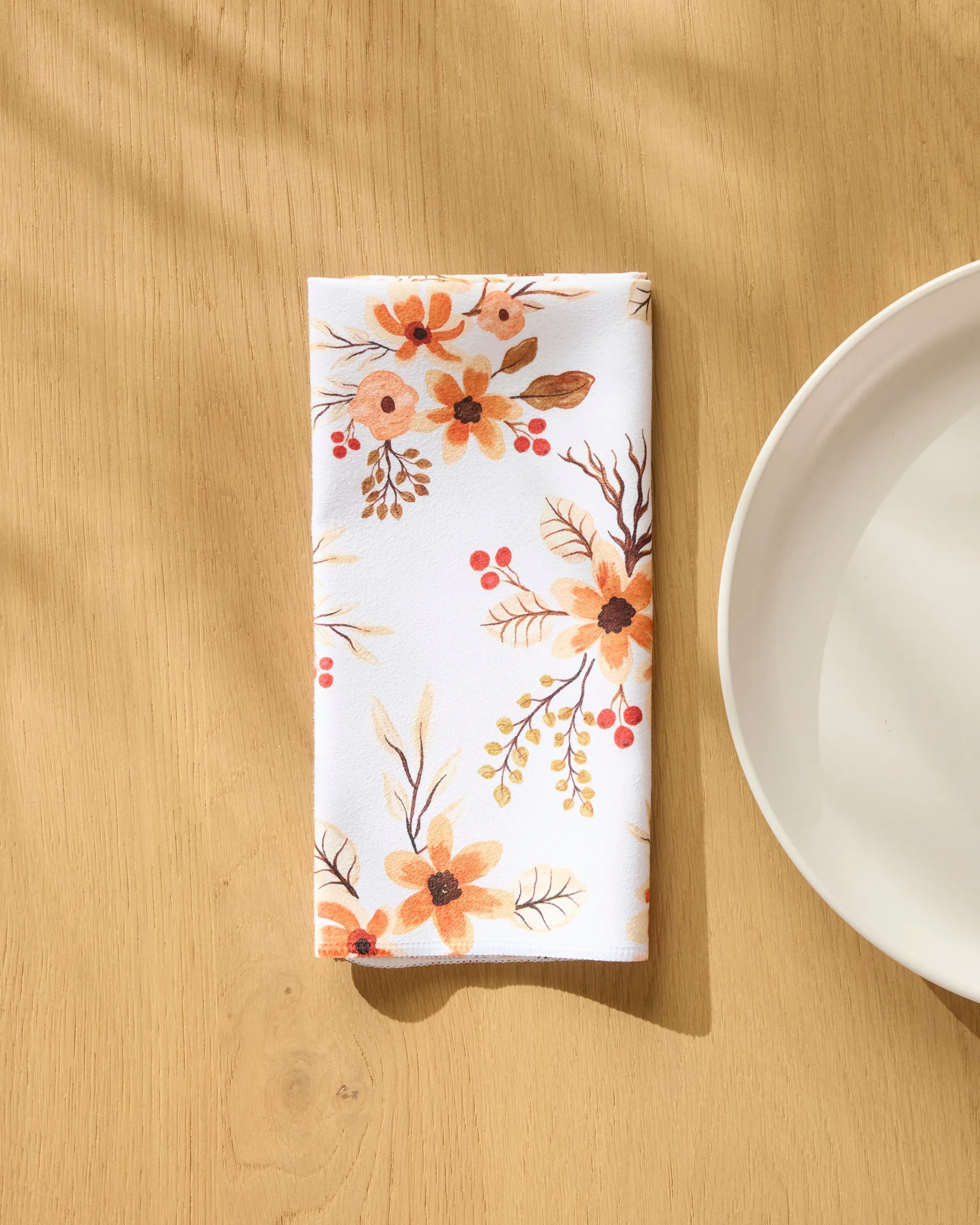 The "In Bloom" eco-friendly napkin by Vaishali Design Studio, featuring orange and yellow floral patterns, rests folded on a light wooden table beside a white plate, bathed in soft natural shadows.