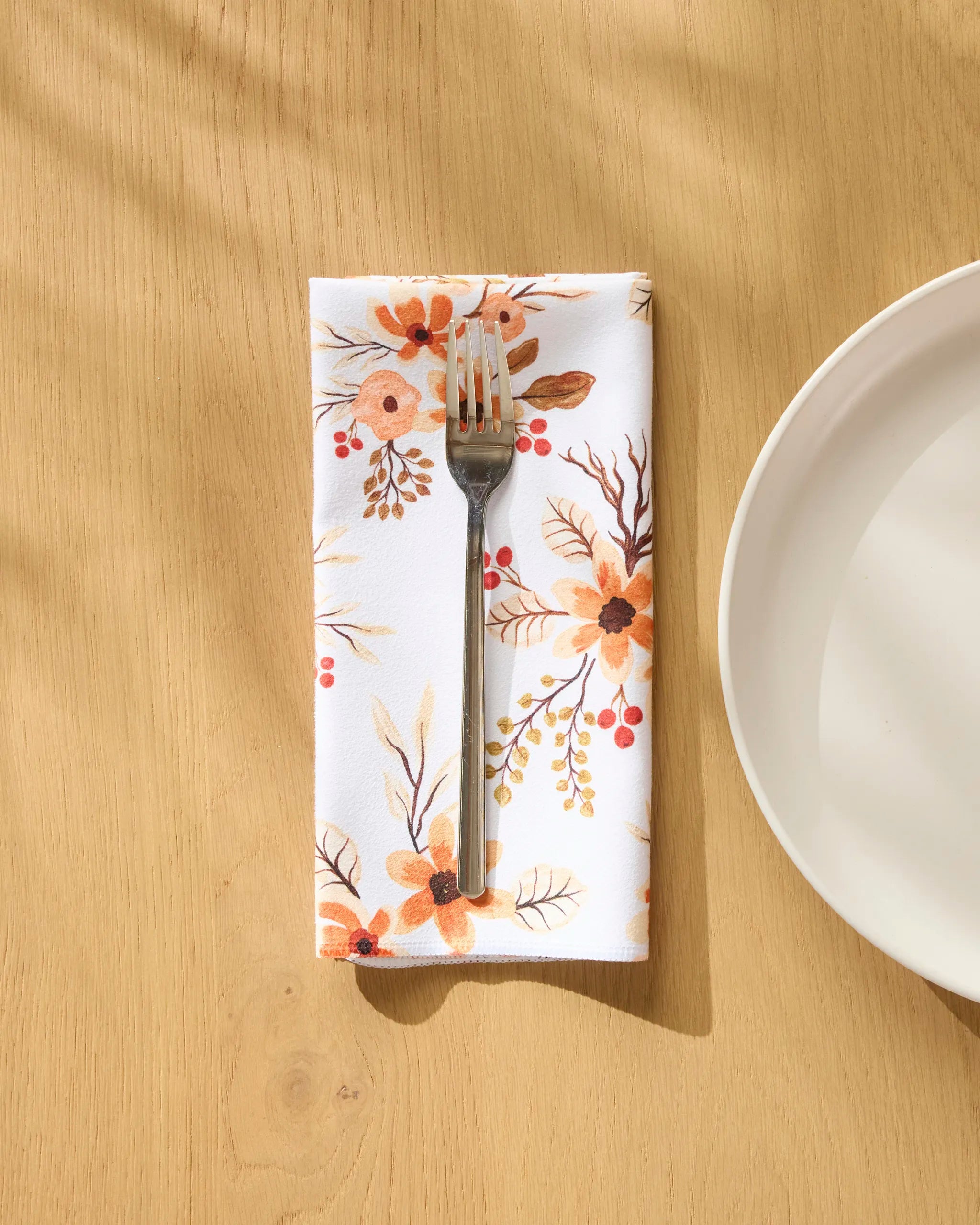 The In Bloom Dinner Napkin Set by Vaishali Design Studio features eco-friendly napkins with orange and beige florals, folded on a light wooden table with a silver fork and white plate. Sunlight creates soft shadows, adding a welcoming touch.