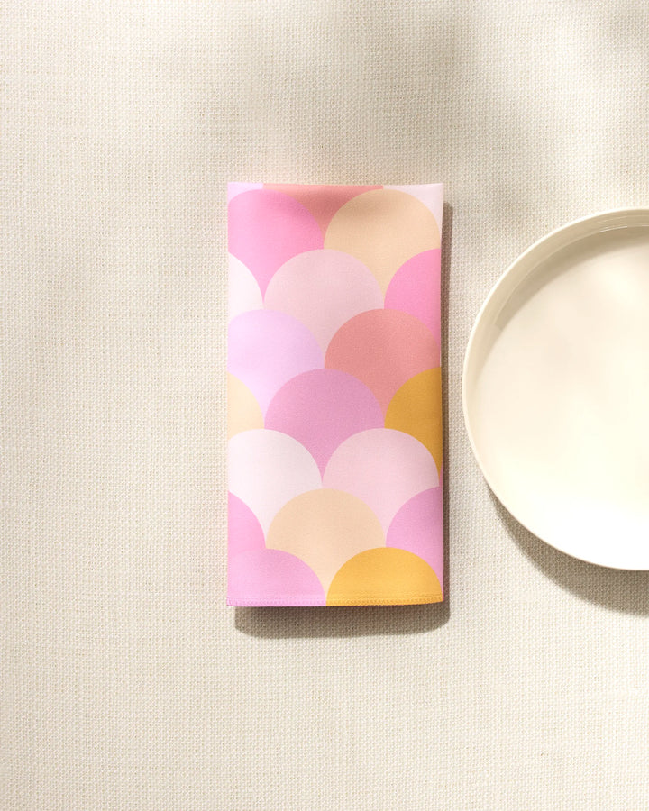 A GEOMETRY Inherit reusable napkin with a pink, purple, and yellow scallop pattern rests beside a plain white plate on a textured, light surface.