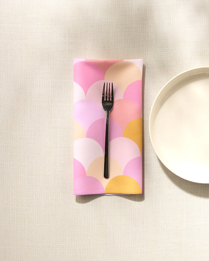 A black fork rests on the GEOMETRY Inherit reusable napkin, featuring pastel pink, lavender, and yellow scallop patterns, next to a plain white plate on a textured light surface.