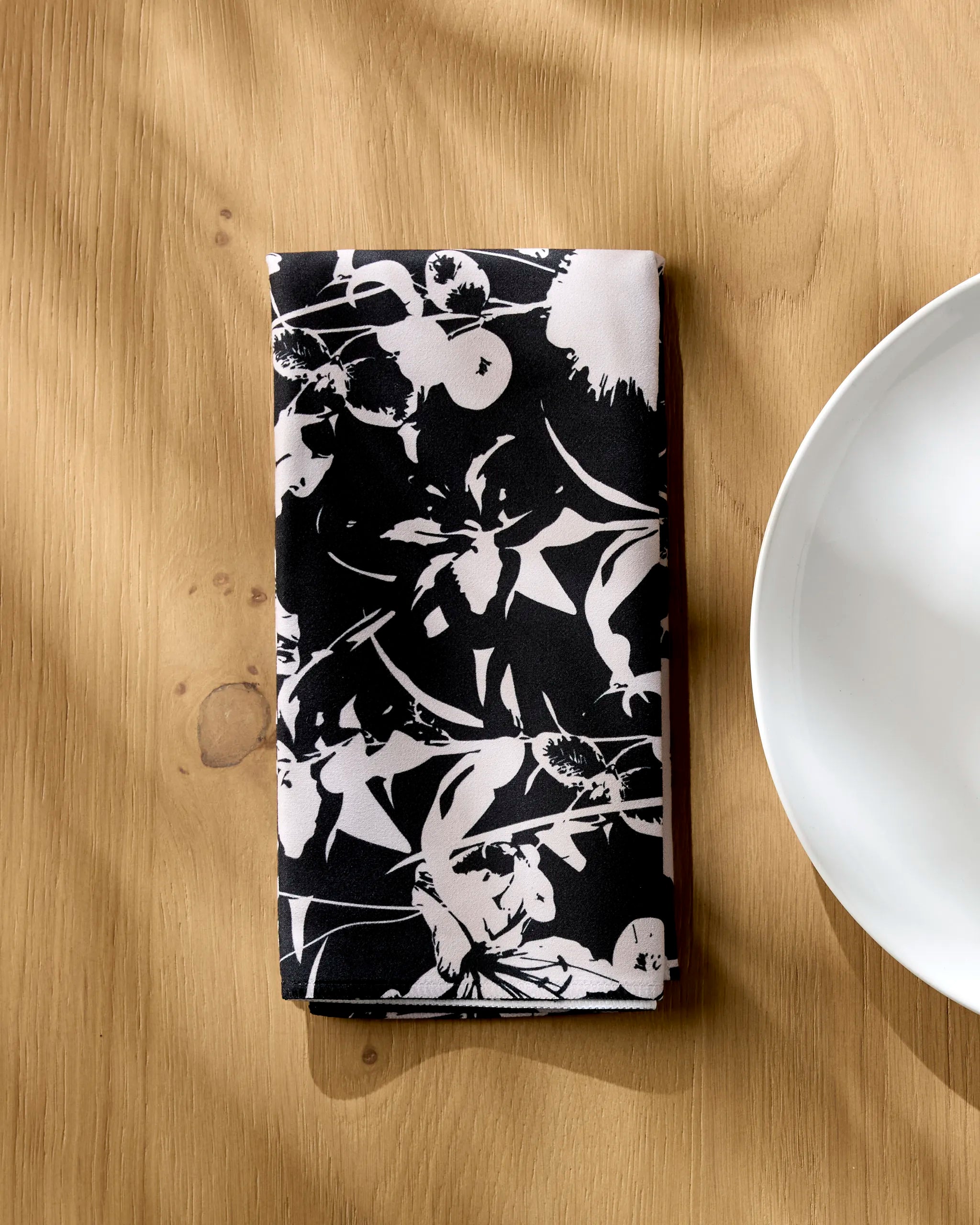 The Geometry Inverted Floral Black reusable napkin, featuring a black and white floral design, is folded on a light wooden table beside a white plate, with soft sunlight casting gentle shadows.