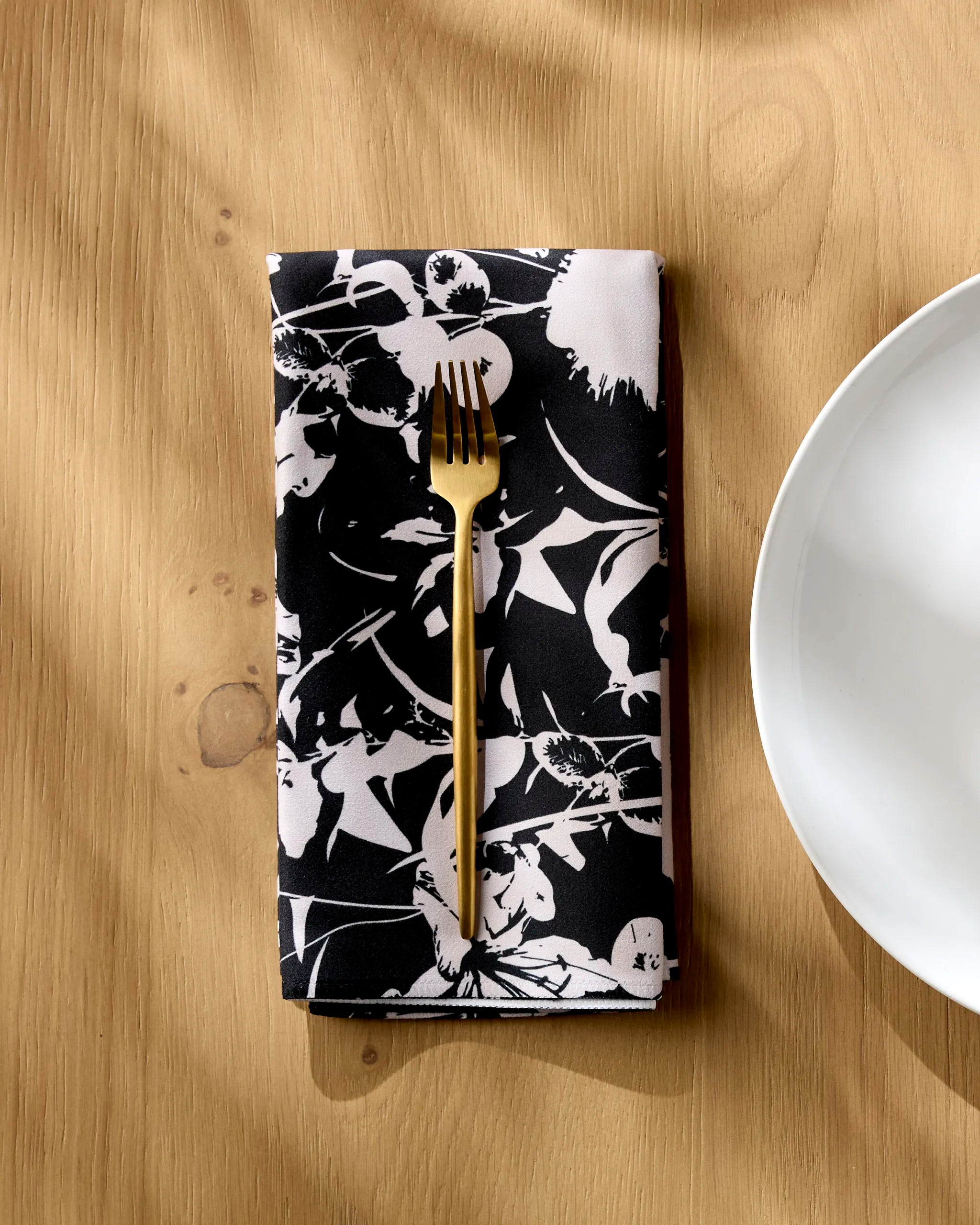 A gold fork rests on a folded Geometry Inverted Floral Black reusable napkin placed on a light wooden table beside a white plate.