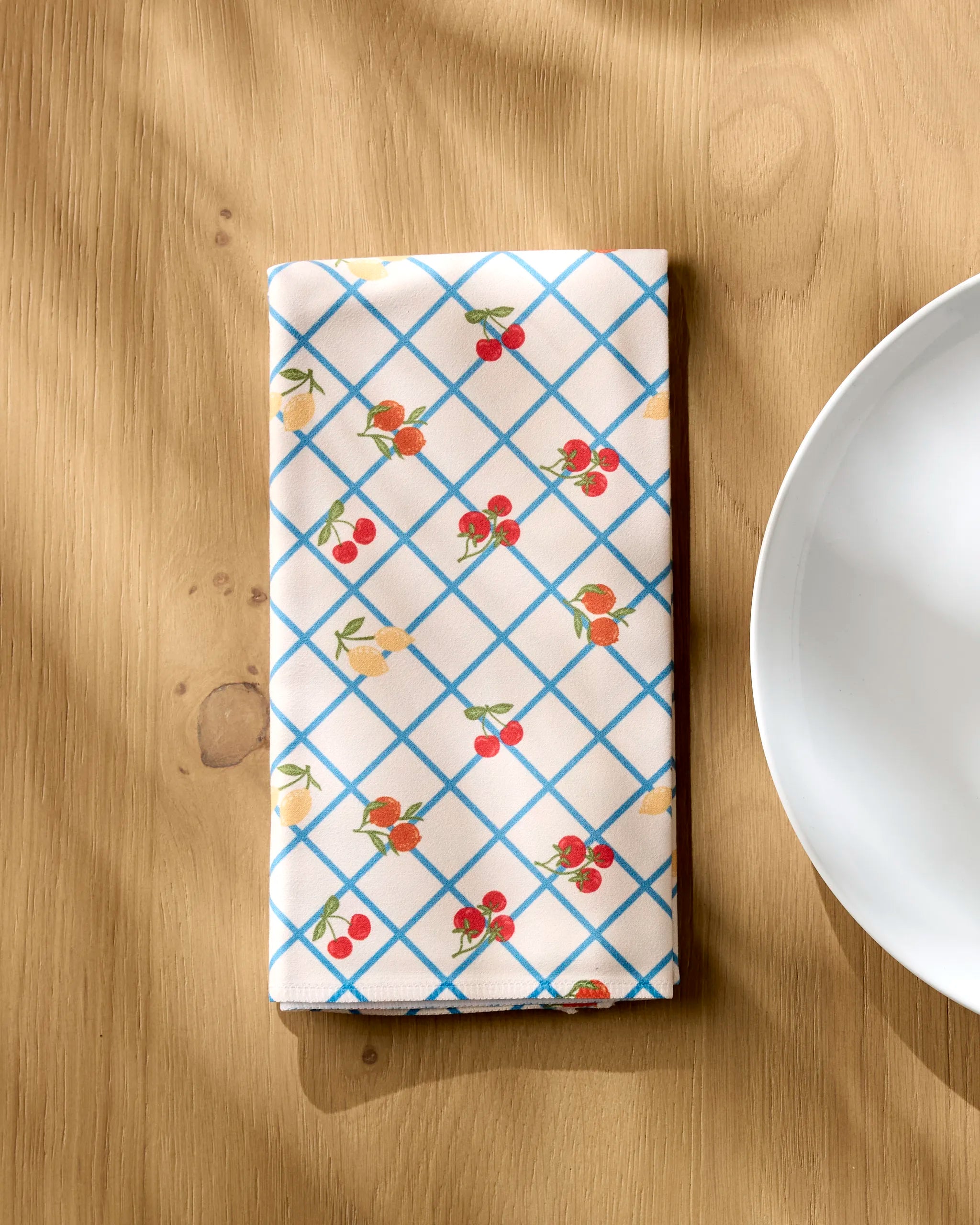 The Geometry Italian Summer Grid recycled dinner napkin, featuring a blue grid and small red and yellow fruits, rests on a wooden table by a white plate, with sunlight casting soft shadows over this absorbent set.