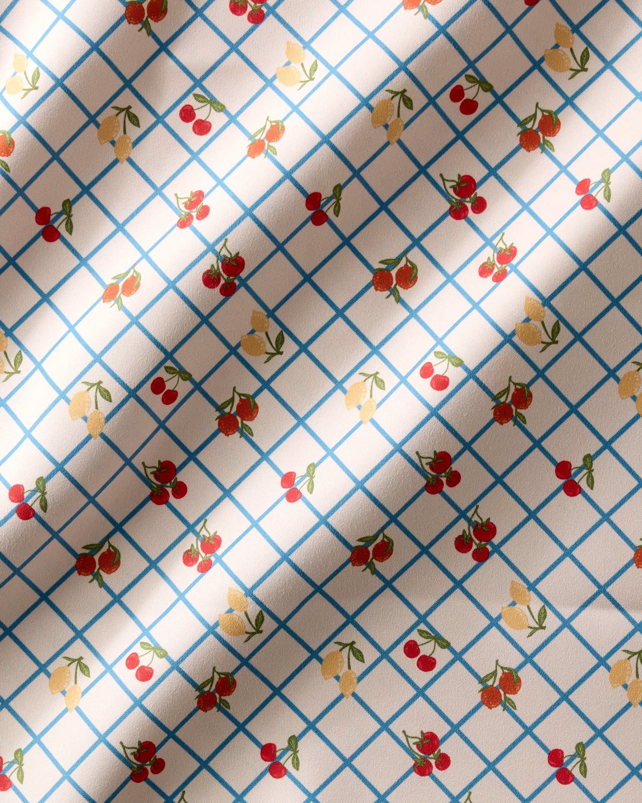 Geometry’s Italian Summer Grid napkins feature a blue diamond pattern adorned with red cherries and yellow strawberries at each intersection, enhanced by sunlight and shadow for a fresh, stylish look.