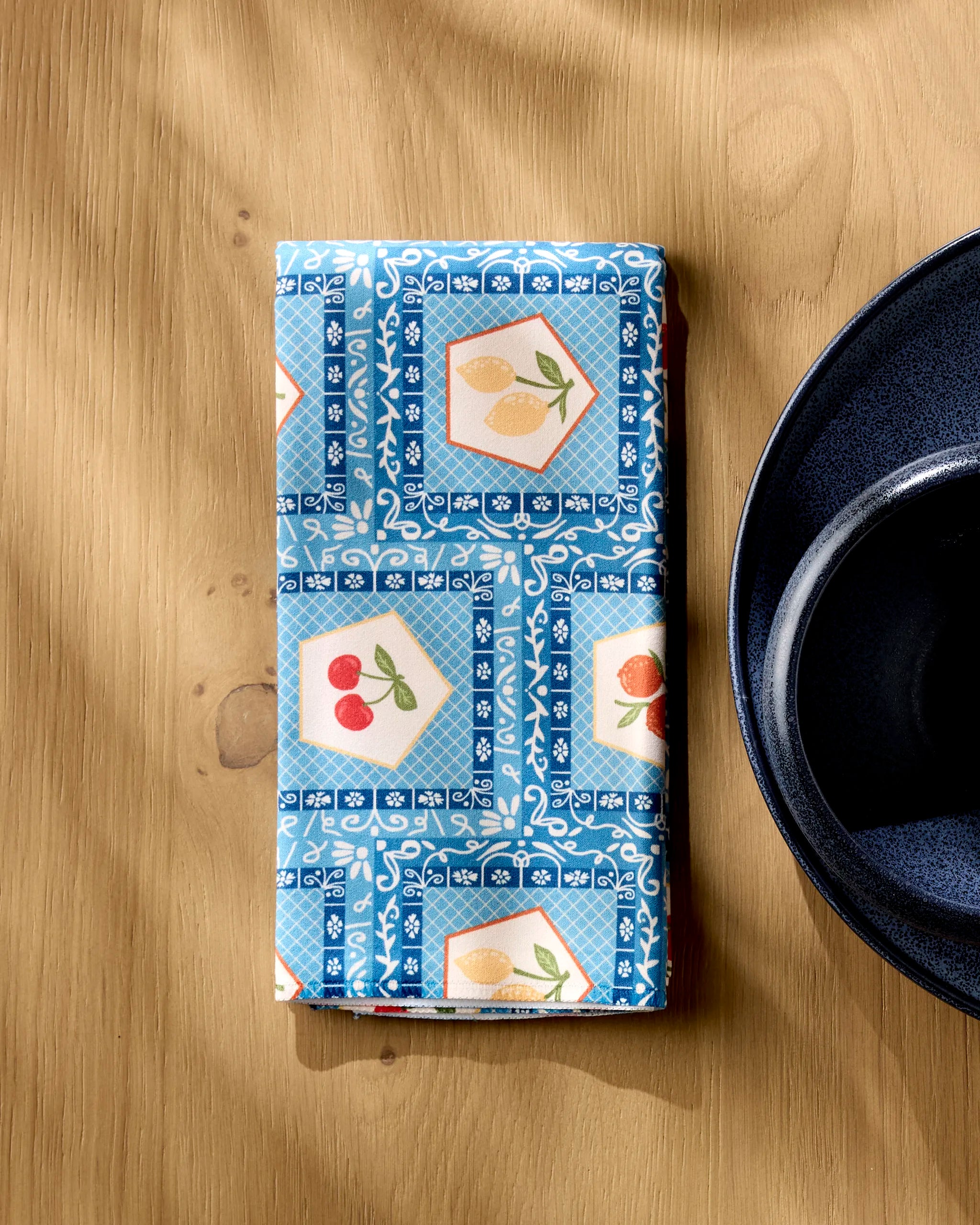 The Geometry "Italian Tiles" recycled dinner napkin, featuring tile-inspired designs in blue, sits folded beside a dark blue bowl on a wooden table.