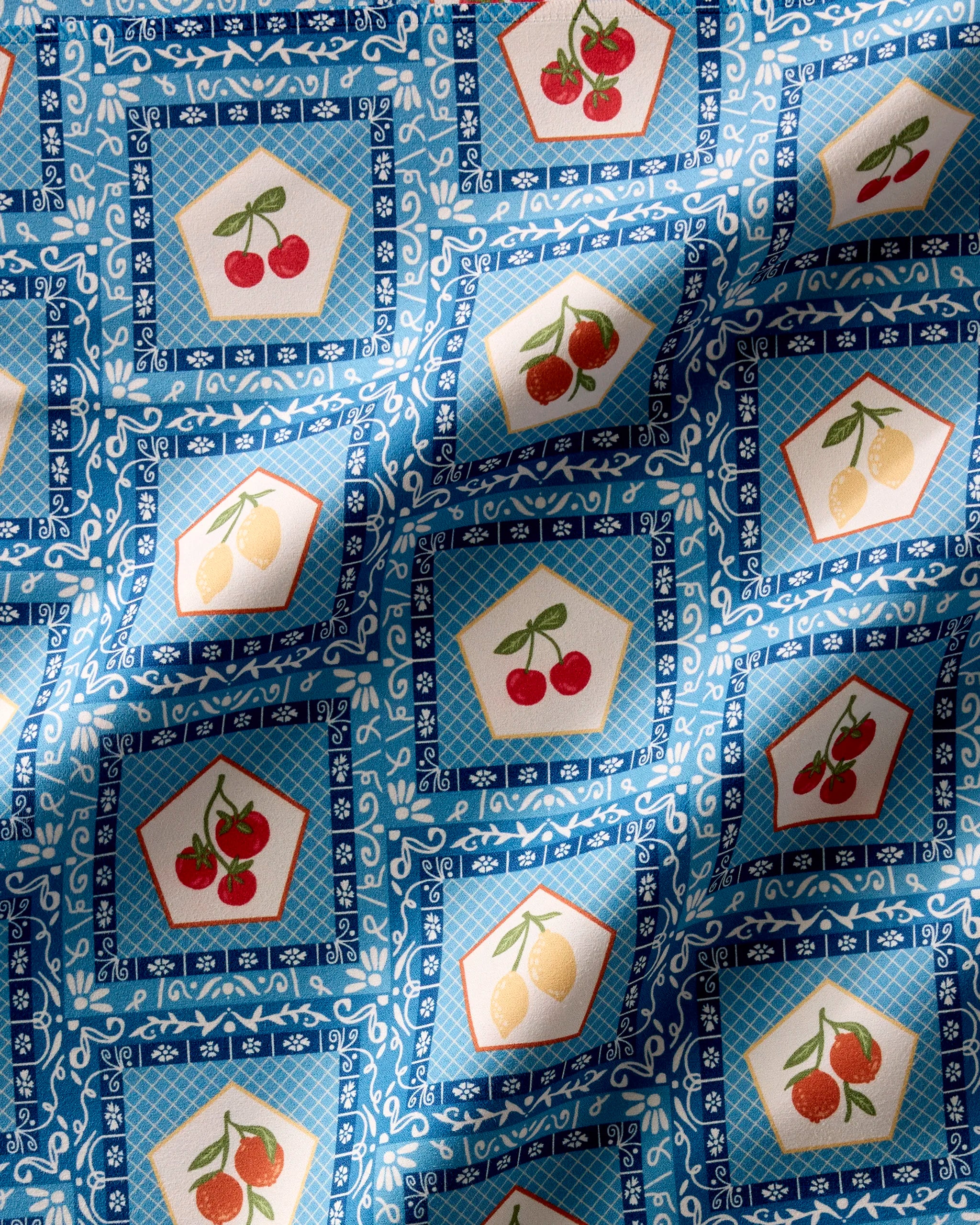 Italian Tiles by Geometry: Blue patterned fabric with white floral designs, showcasing pentagon-shaped frames of cherries, lemons, and strawberries in a grid. Slightly wrinkled—ideal for absorbent cloth napkins or a recycled Dinner Napkin Set.