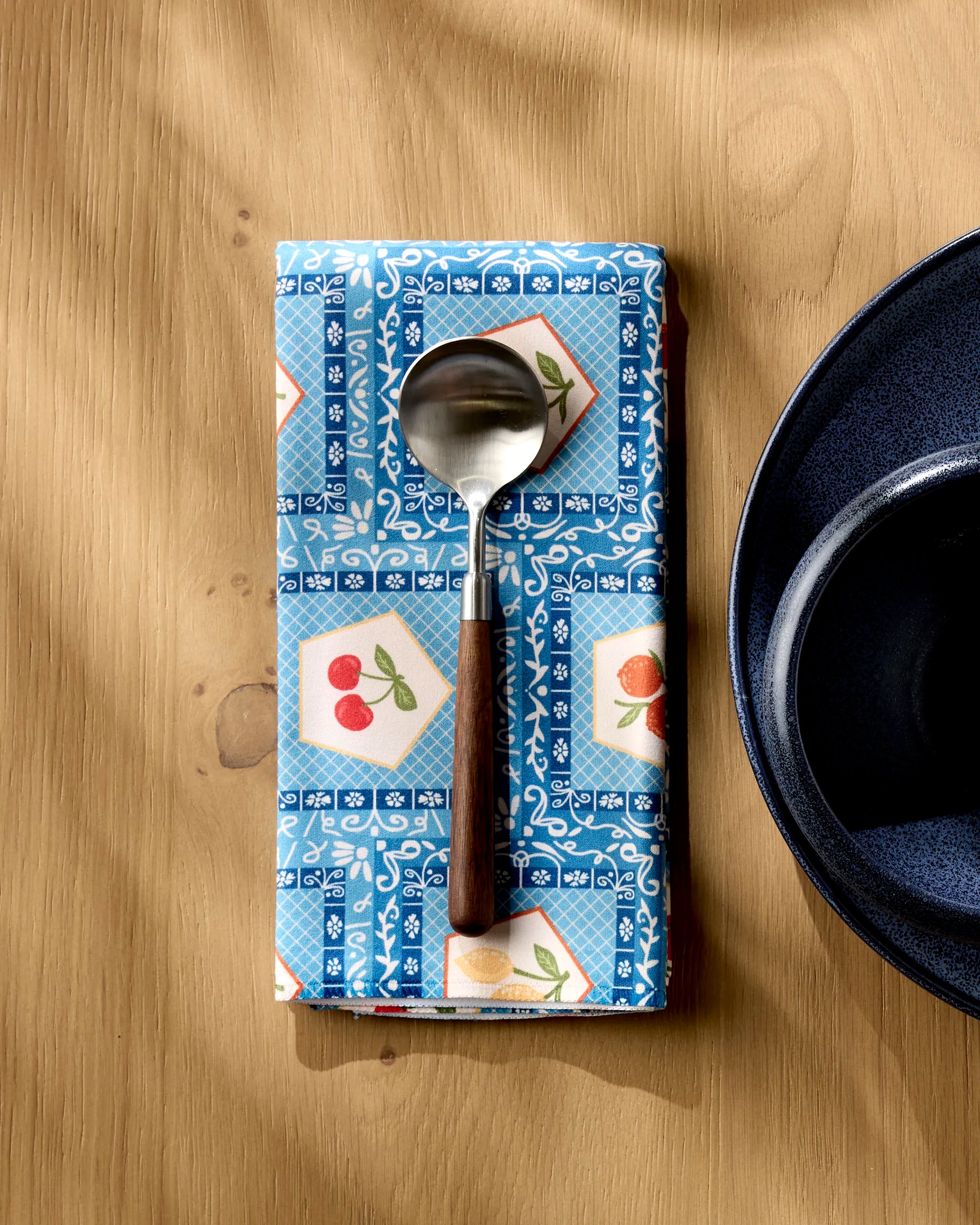 A Geometry Italian Tiles absorbent cloth napkin with colorful fruit designs sits on a wooden table, supporting a spoon with a wooden handle next to a dark blue bowl.