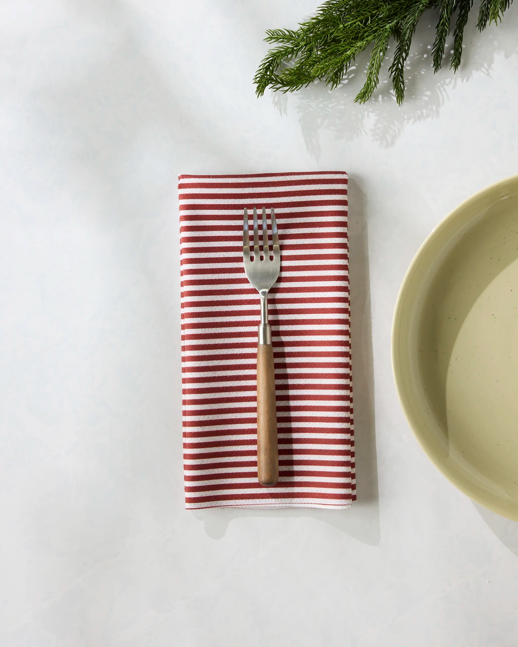 A Kringle reusable napkin by Geometry is neatly folded with a fork on top, placed next to a beige plate on a white surface, accented with green pine branches in the corner.