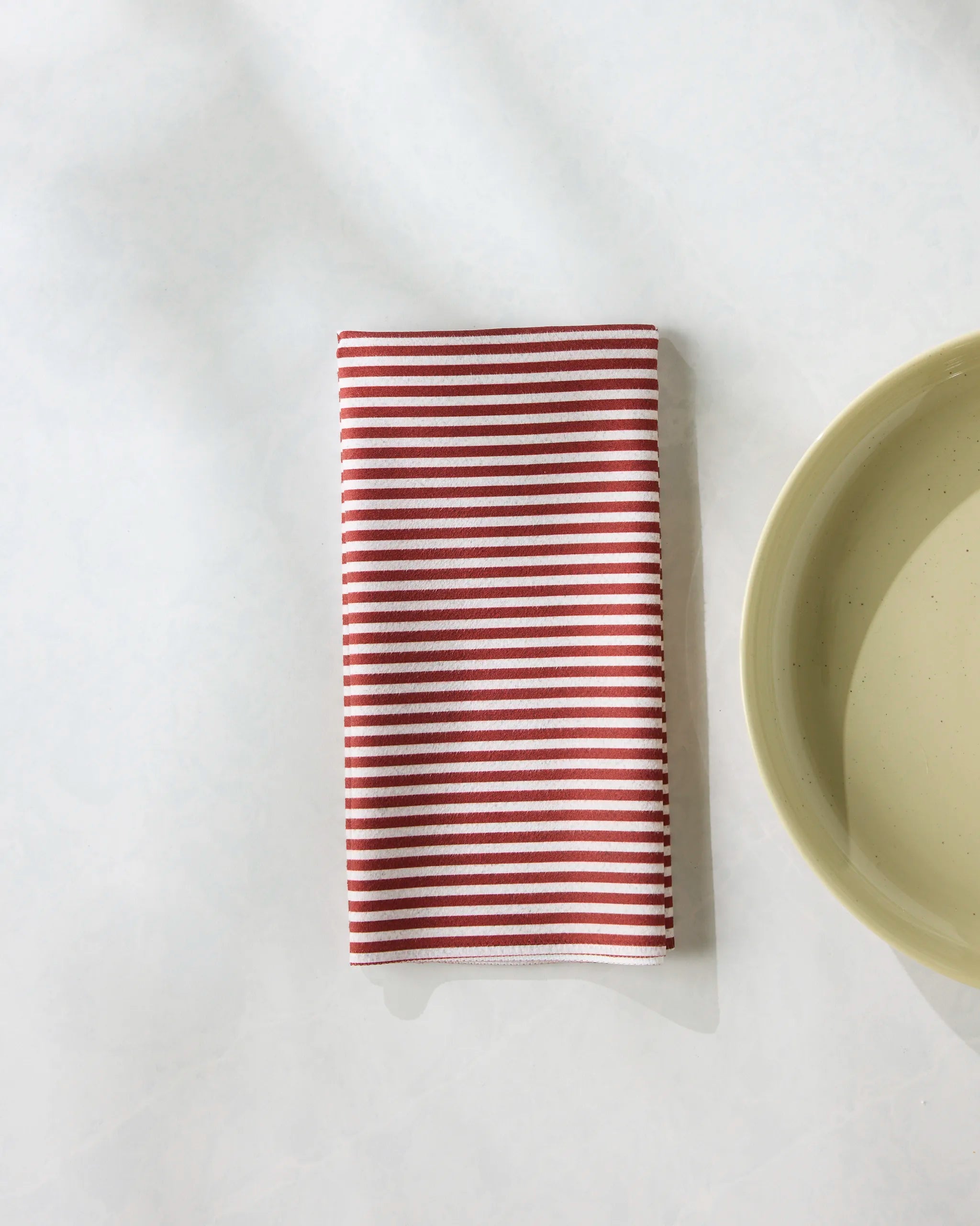 A neatly folded Geometry Kringle reusable napkin with red and white stripes rests on a white surface beside a light beige plate.