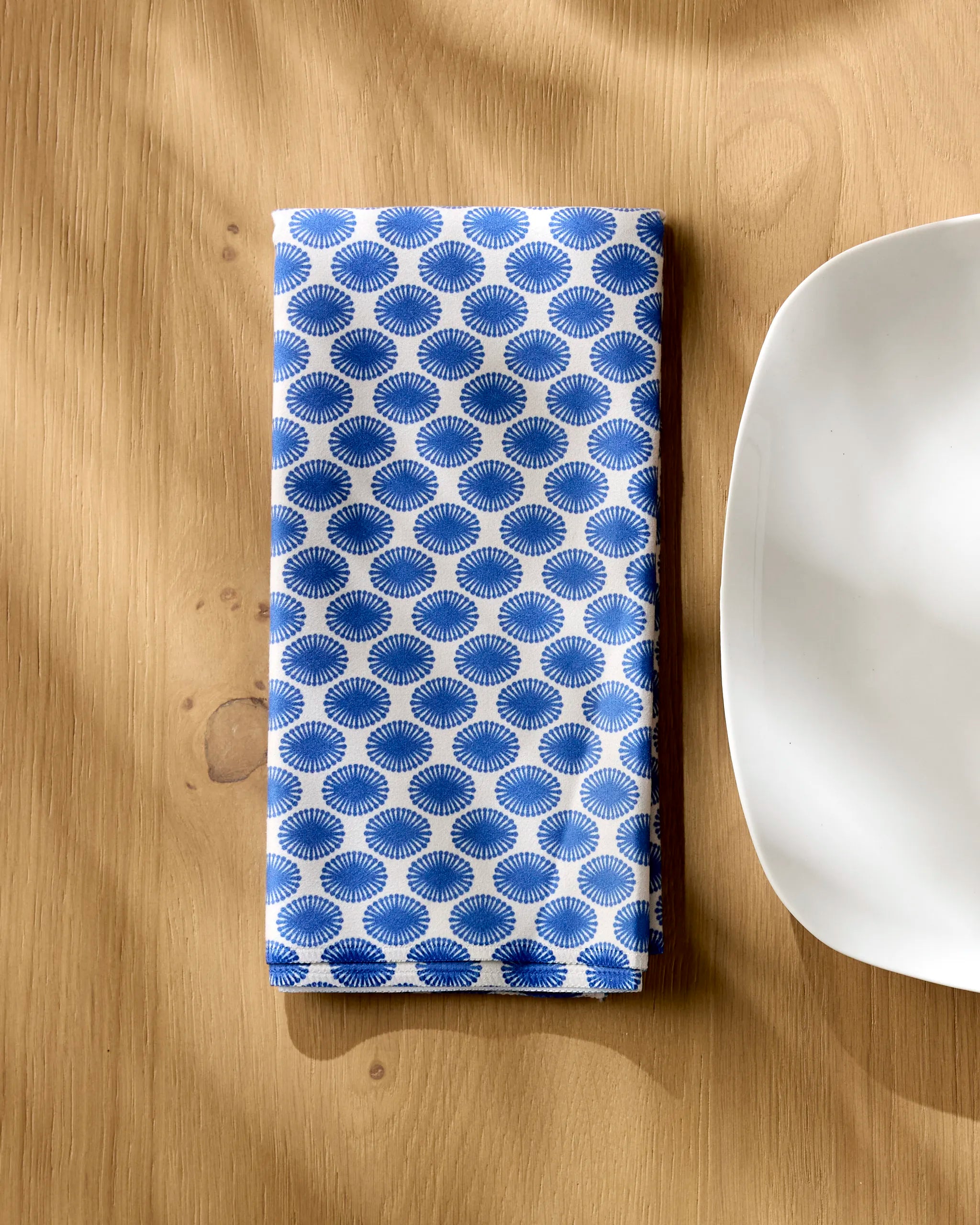 The Lagoon reusable cloth napkin by Geometry, featuring a blue and white pattern, is folded on a light wood table beside the corner of a white square plate, with soft shadows across the scene.